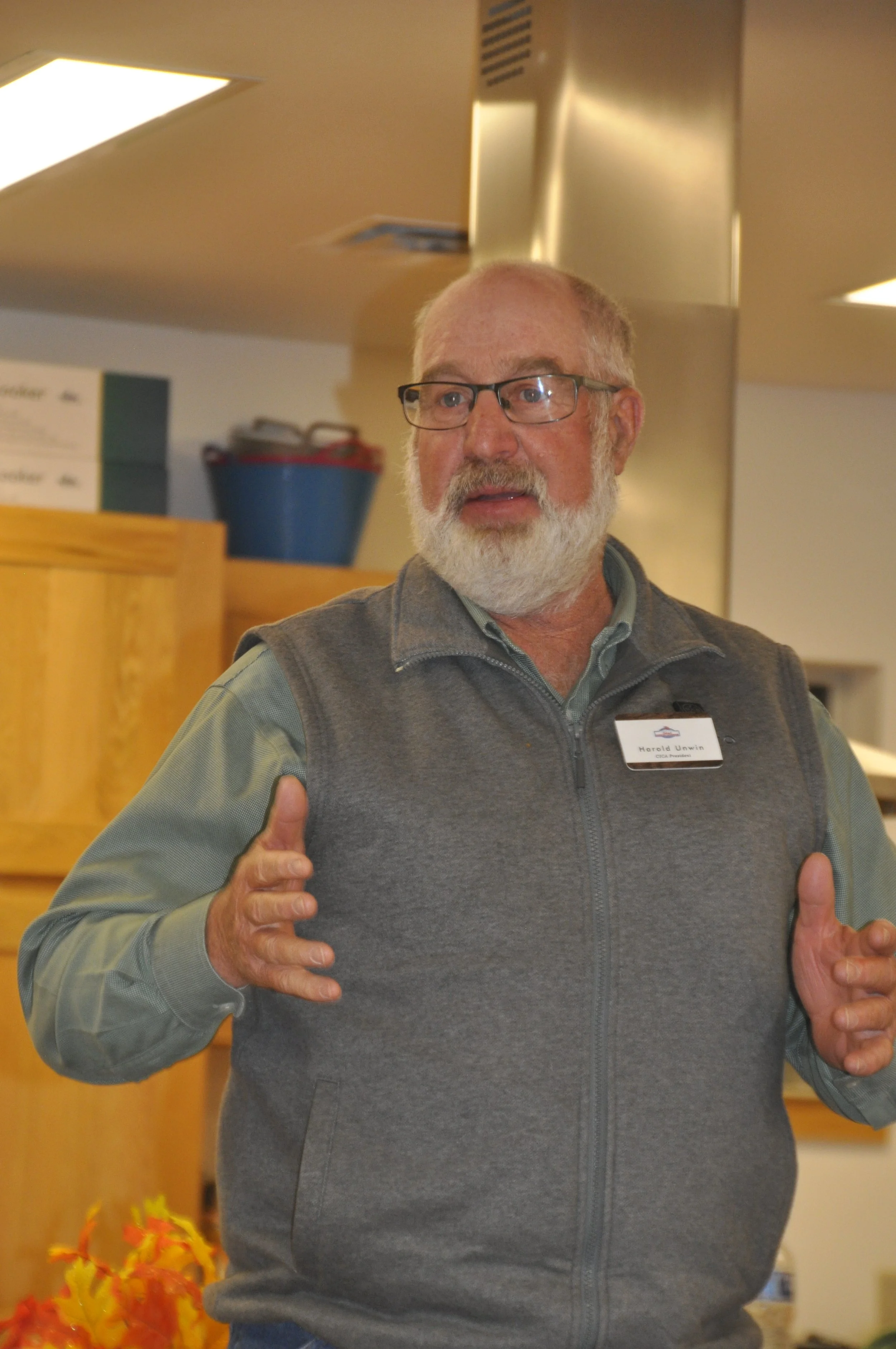 An elderly man with glasses and a beard, wearing a gray vest over a light shirt, standing indoors. He is giving two thumbs up and appears to be speaking or explaining something, with a name tag that reads 'Harold Unwin'.