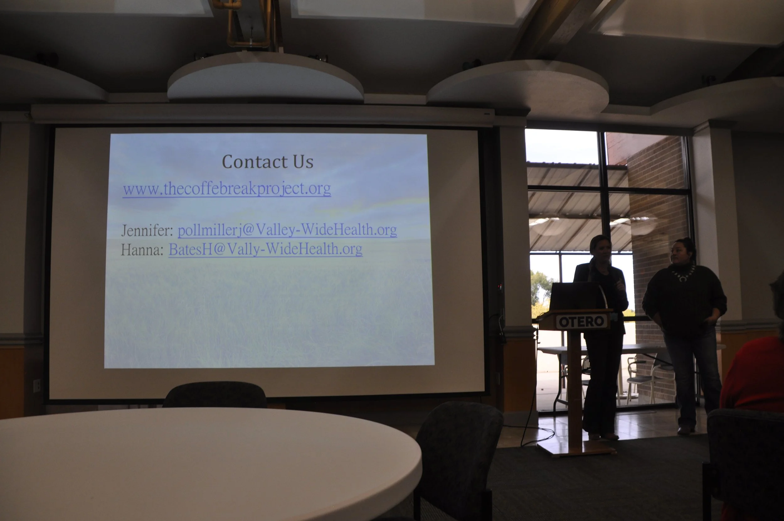 A presentation slide on a large screen providing contact information for a project, with names and email addresses, in a room with large windows showing a brick building outside, and two women standing near a podium labeled 'OTERO'.