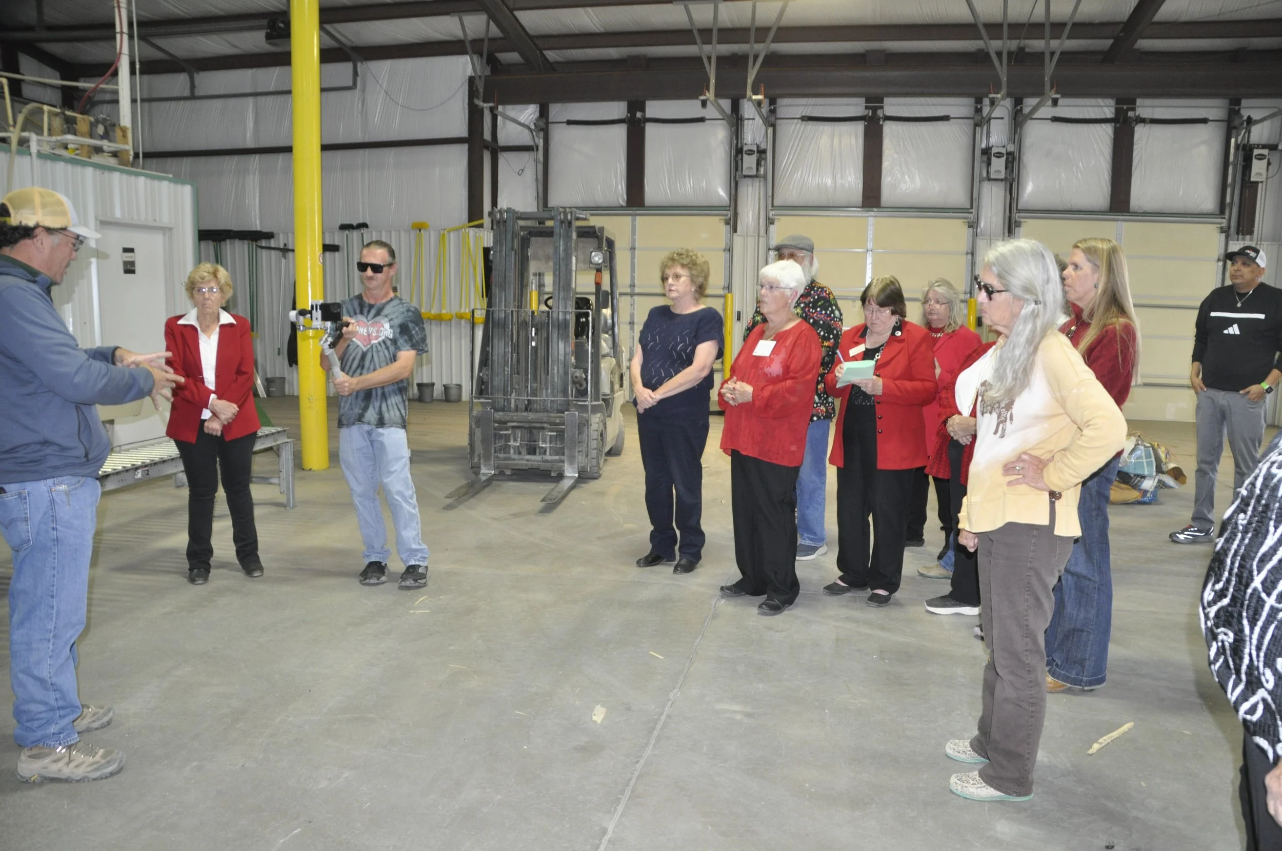 Group of people standing inside a warehouse, listening to a man in a blue shirt who is speaking. The group is standing on a concrete floor near a forklift and yellow pillars, with a corrugated metal wall and garage doors in the background.