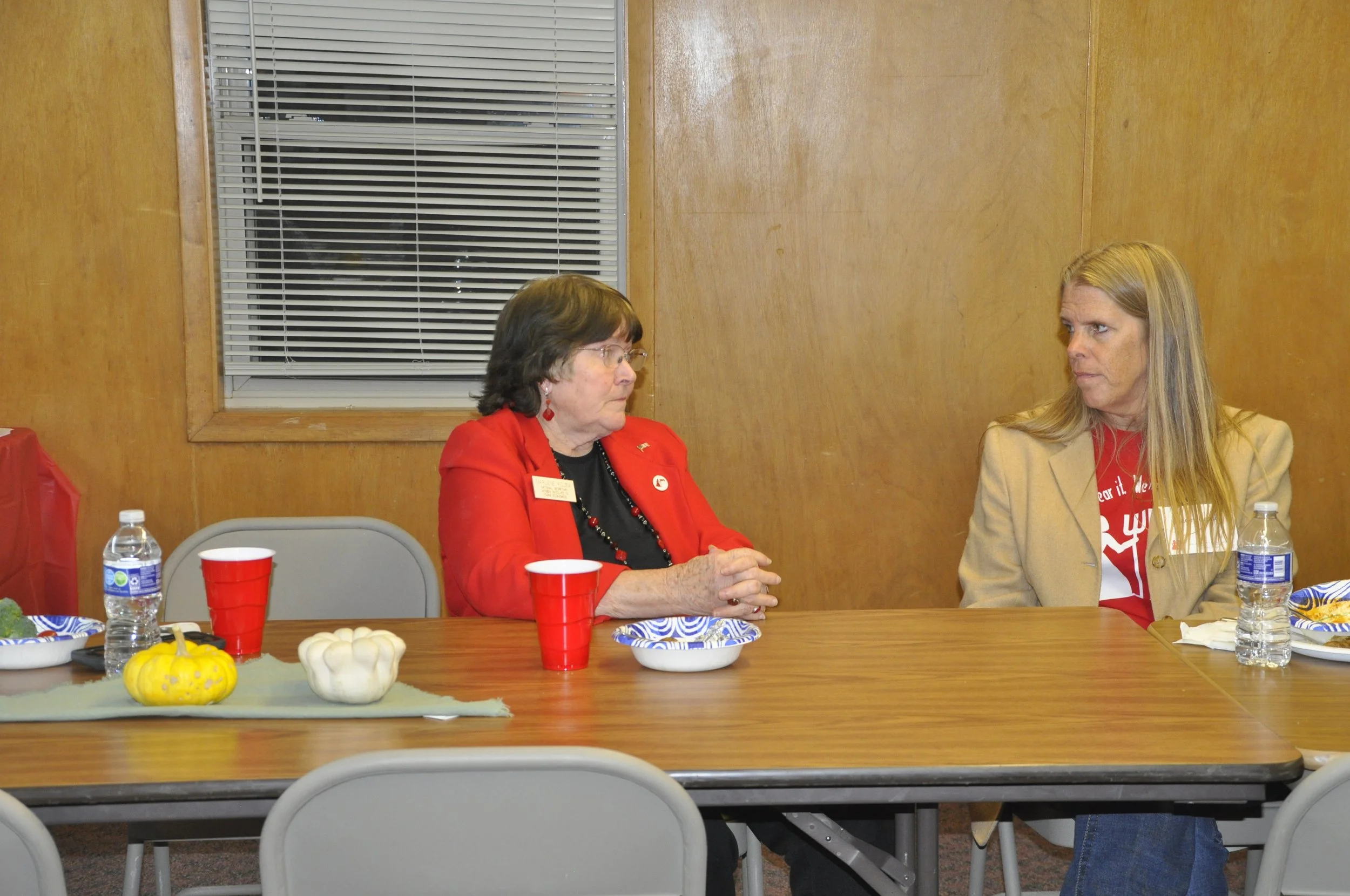 Two women sitting at a table during an indoor gathering or meeting. The woman on the left is older, wearing glasses, a red blazer, and earrings. The woman on the right has long blonde hair, wearing a beige blazer and a red shirt. The table has water 