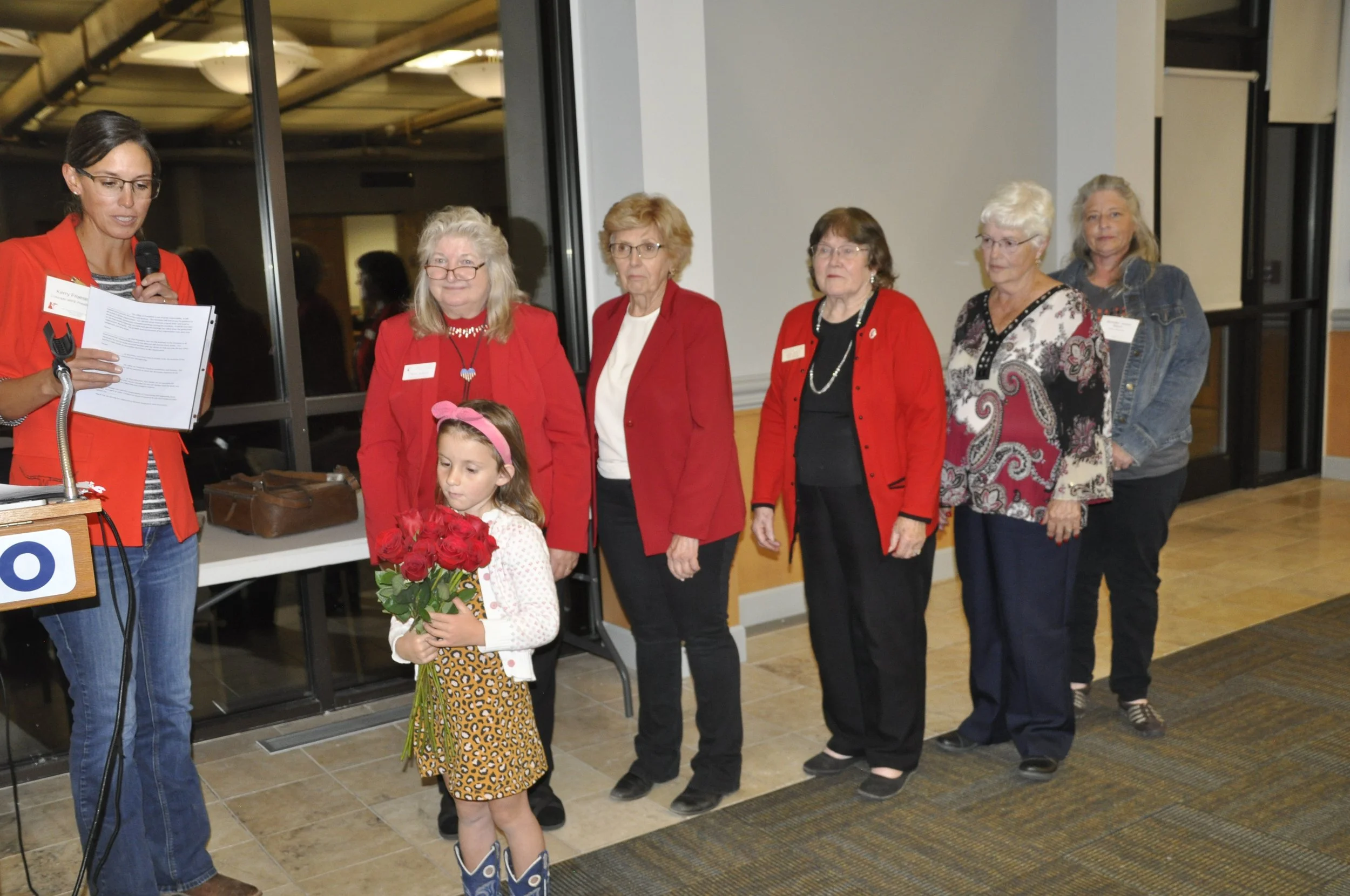 A woman speaking into a microphone reads from a paper at a recognition event. She stands beside a young girl holding a bouquet of red roses. The girl wears a leopard-print skirt and pink hairband. Four older women stand in a line behind them, all dre