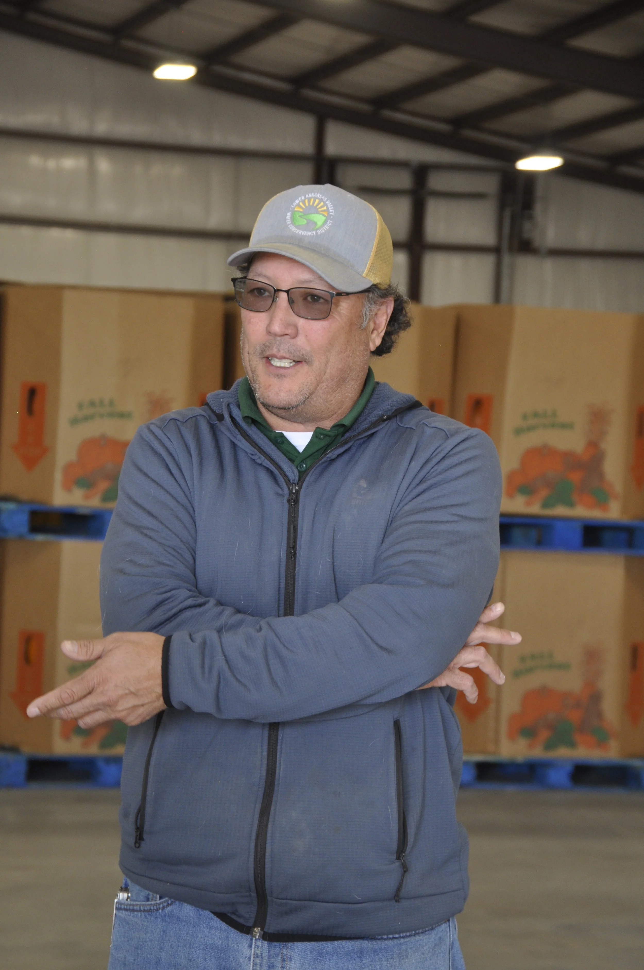 A man wearing glasses, a gray baseball cap, gray jacket, and jeans standing inside a warehouse with cardboard boxes in the background.