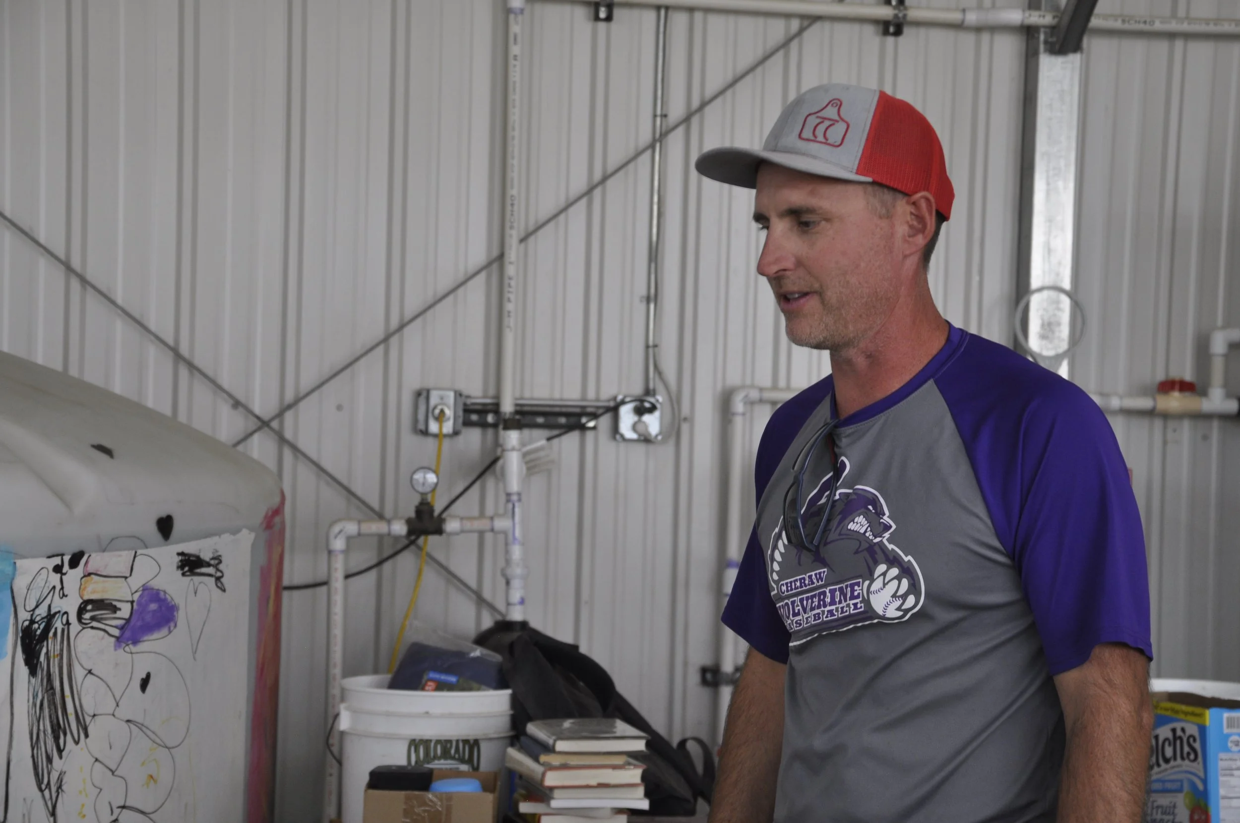 A man wearing a gray and red baseball cap and a purple sports shirt with the Cheraw Wolverines logo is looking to the side inside a garage or workshop, standing next to a colorful drawing on a whiteboard or poster. There are shelves with books, a buc