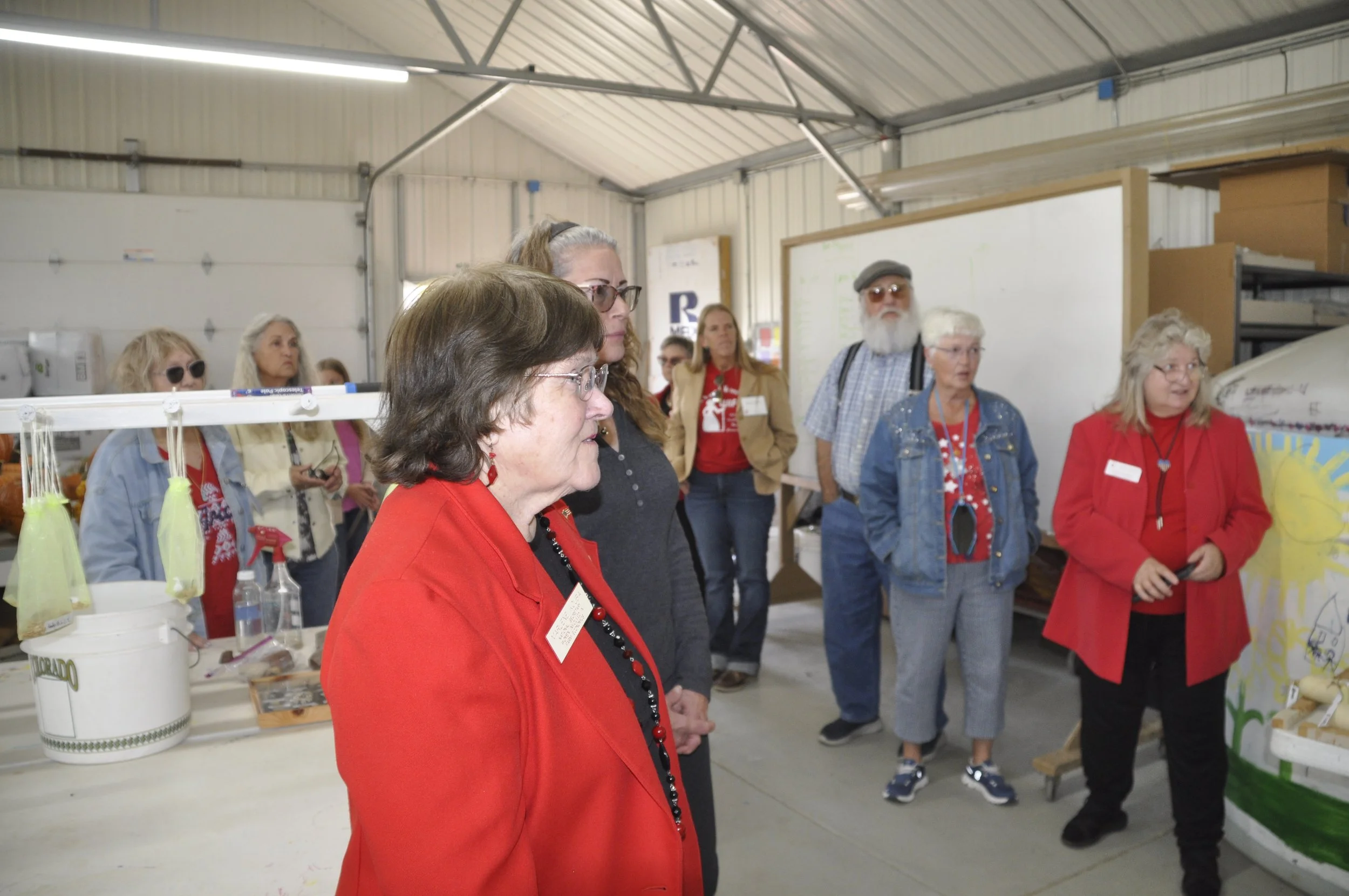 A group of people, mostly women, standing inside a warehouse or workshop with metal roofing. Some are observing a display or presentation, with products and informational materials on tables nearby.