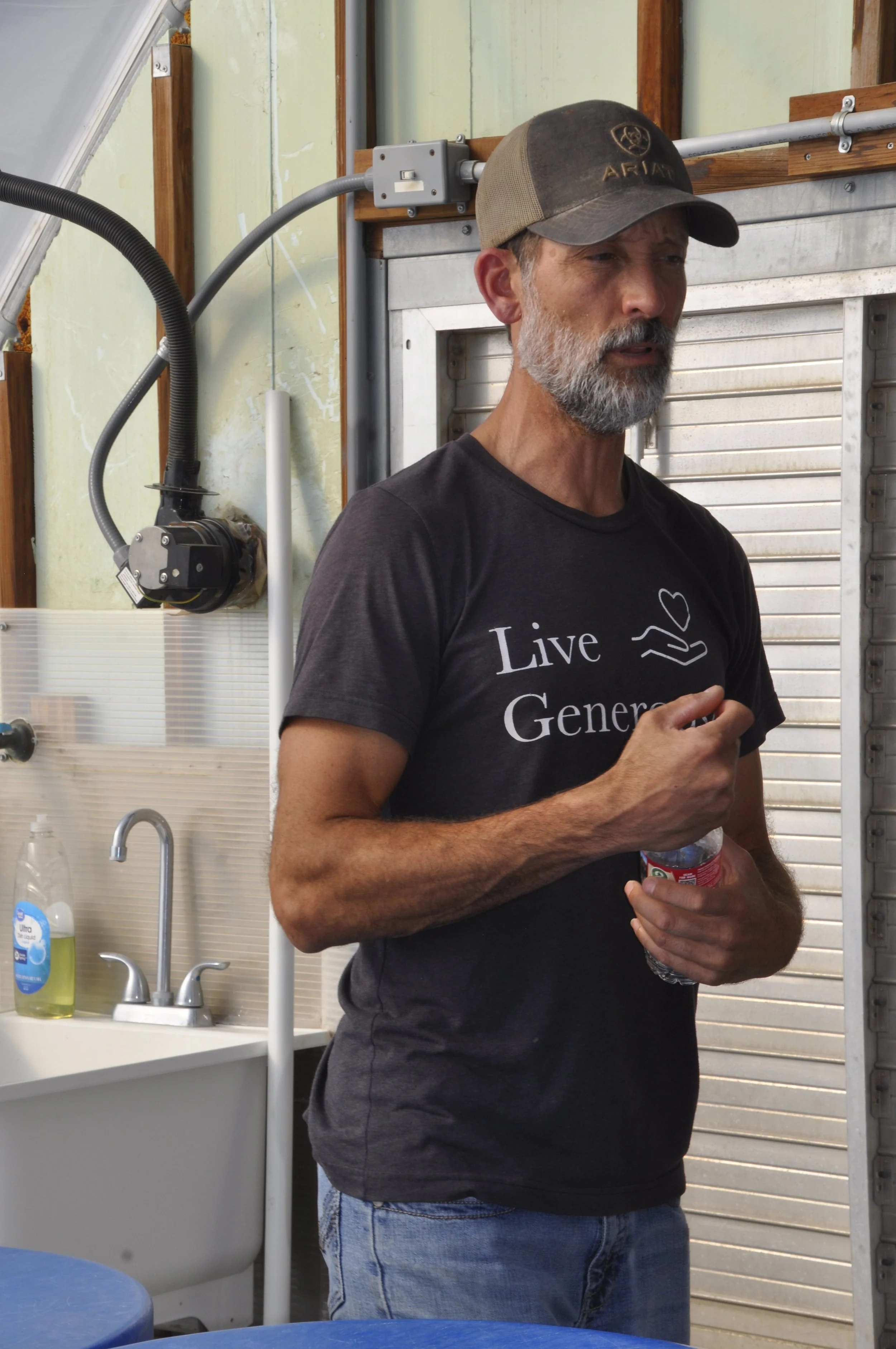 A man with gray hair and beard wearing a gray baseball cap and a black T-shirt with the words "Live Generously" in white, standing in a kitchen or workshop area, holding a small plastic bottle in his left hand. The background includes a stainless ste