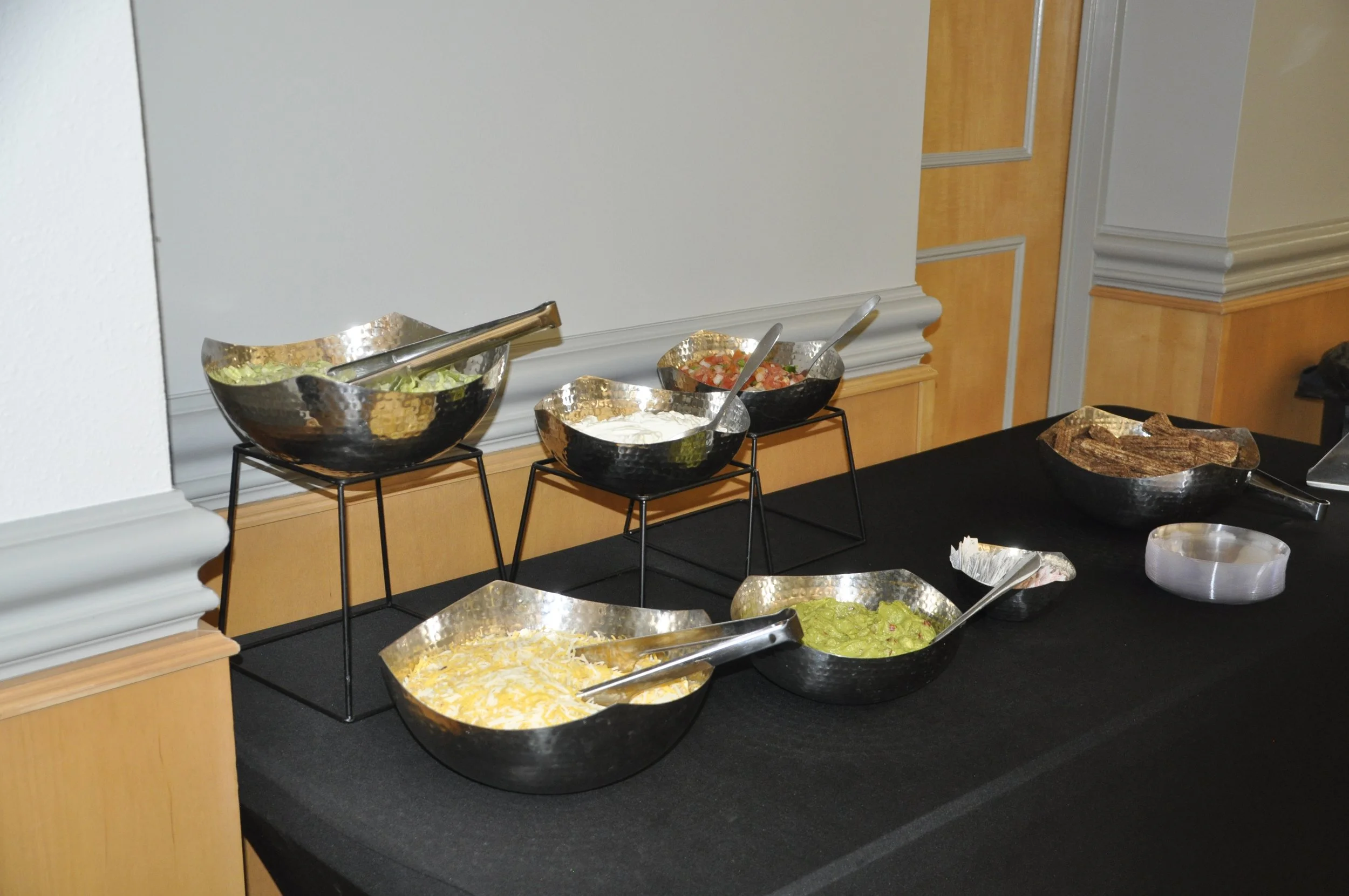 Buffet table with bowls of salad, salsa, and creamy dip on black tablecloth in a room with wood paneling and beige walls.