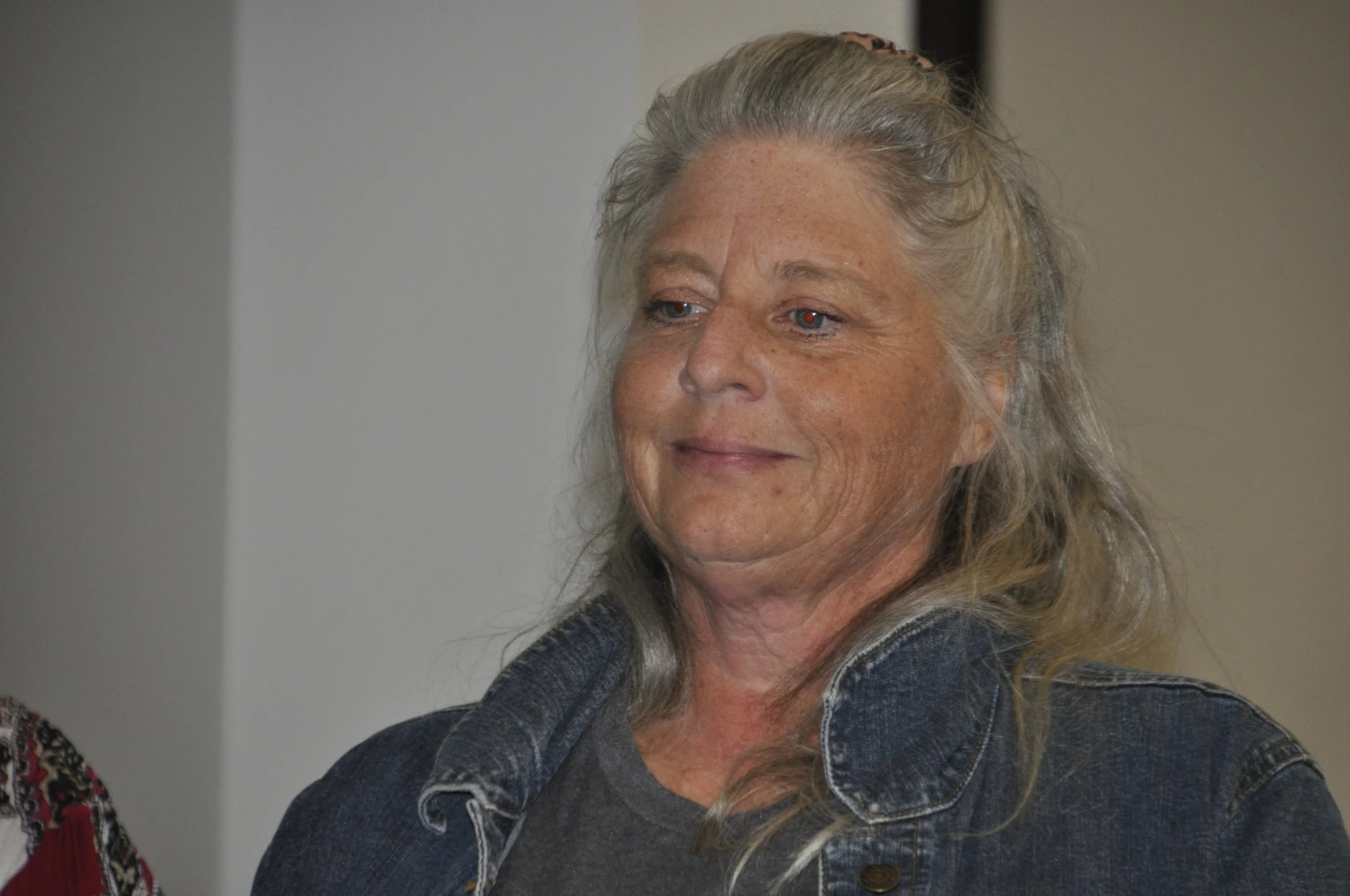 Close-up of a middle-aged woman with long, wavy gray hair, wearing a denim jacket, looking slightly to the right with a neutral expression.