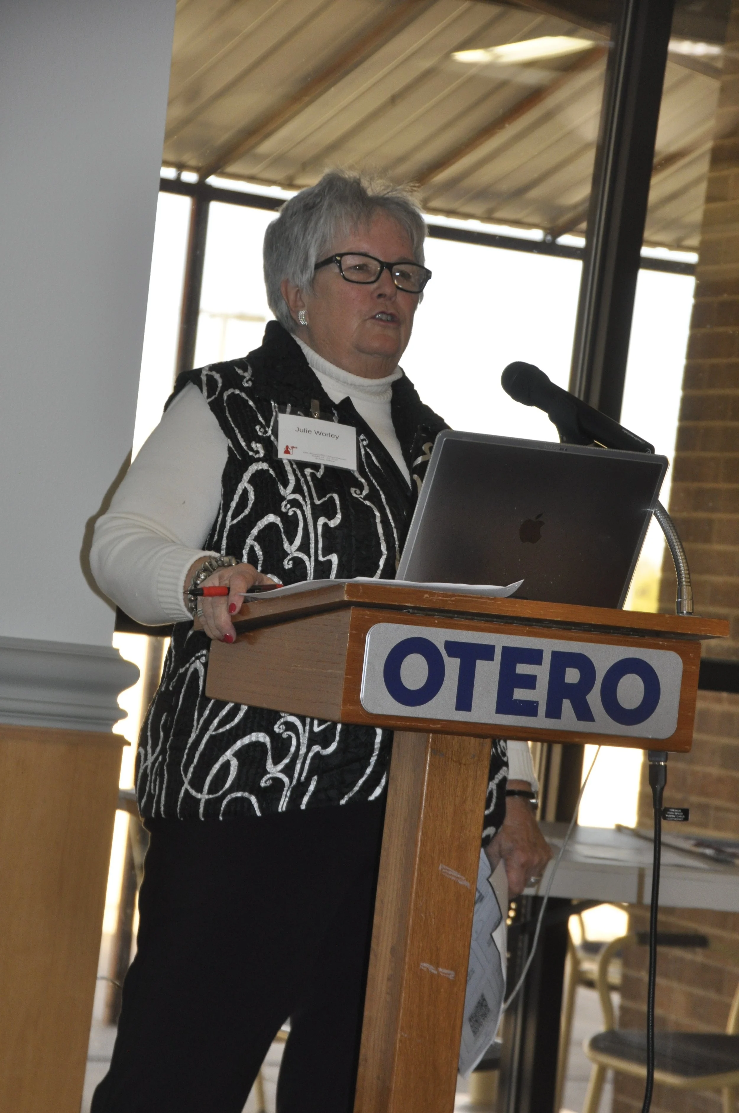 A woman with gray hair and glasses speaking at a podium with a microphone. She is wearing a black and white patterned vest over a white shirt and has a name tag that reads 'Julie Worthey.' The podium has a sign with the word 'OTERO.' She is inside a 