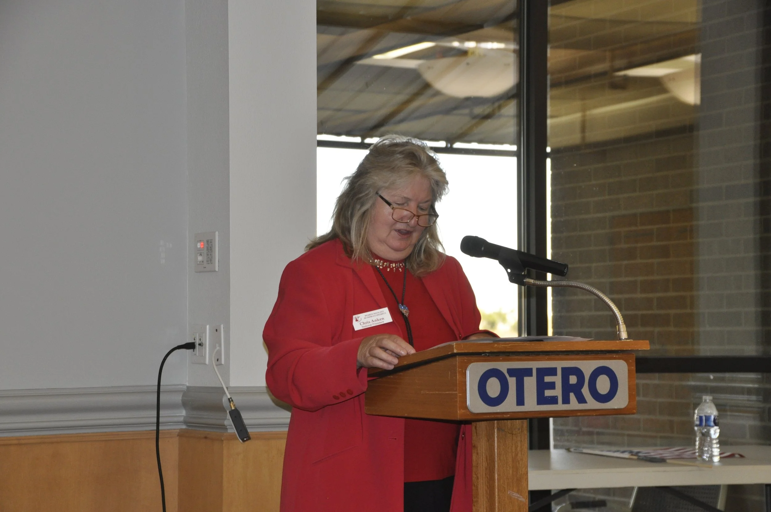 A woman with gray hair wearing glasses and a red blazer standing at a podium with a microphone, reading or speaking. The podium has a sign that says 'OTERO.' Behind her, there are large windows with a view of a brick building outside, and a water bot