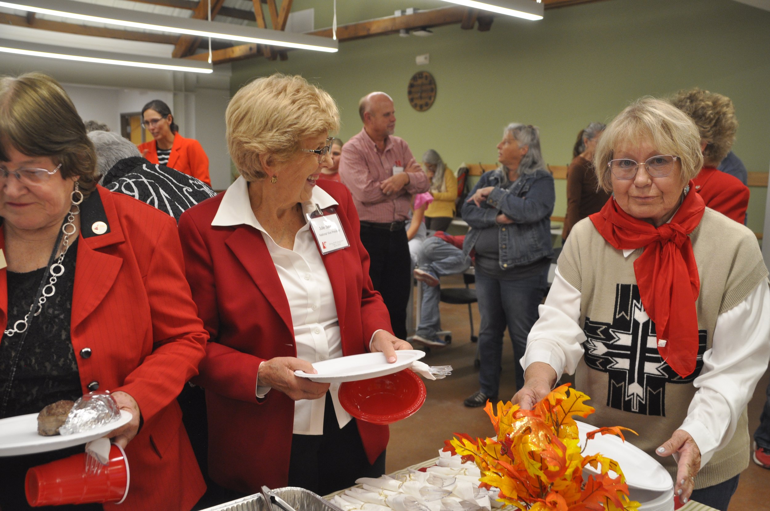 Group of older adults gathered at a table during a social event, some holding plates with food, decorations with autumn leaves on the table, and people in the background engaging in conversations.