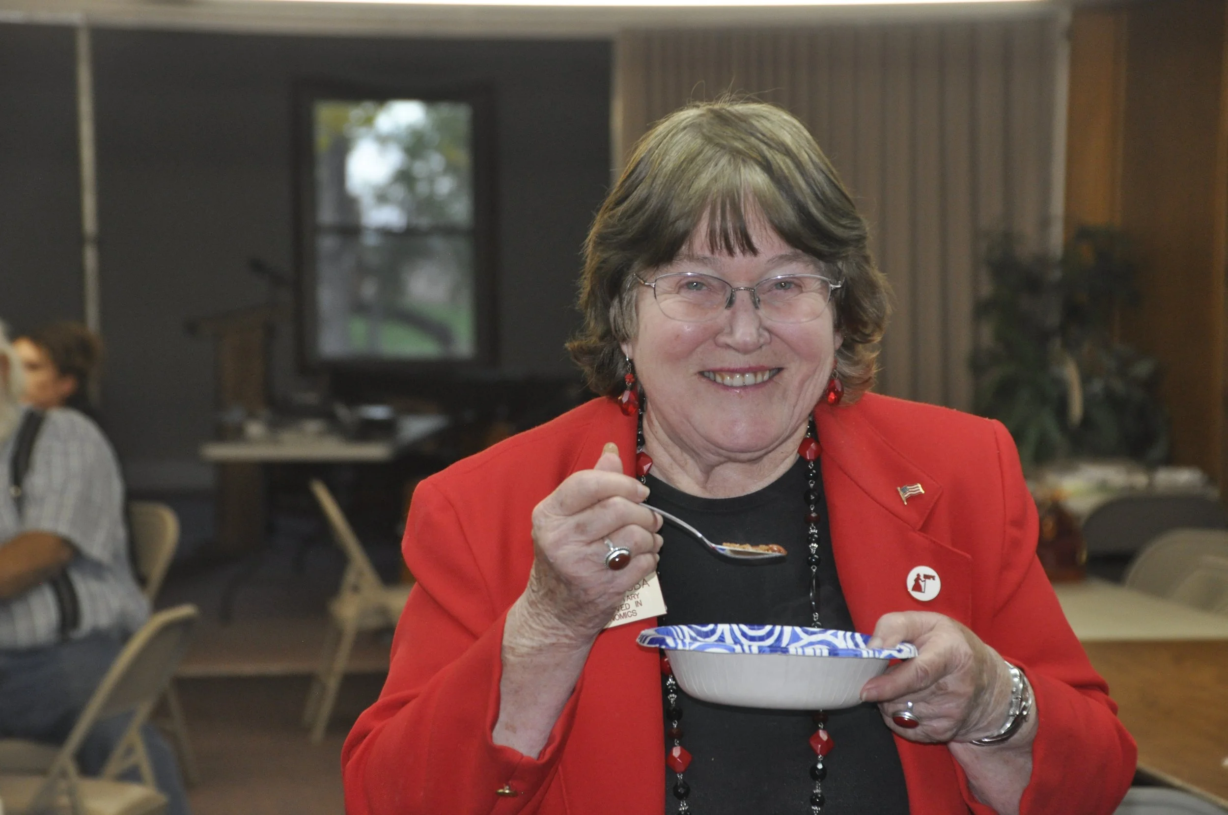 A smiling woman with glasses and earrings, wearing a red blazer with a patriotic pin, eating food from a bowl at an indoor event.