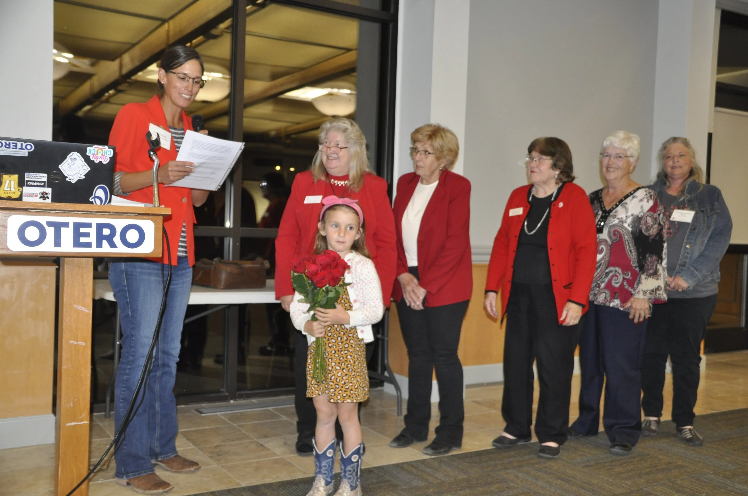 A woman in a red blazer stands at a podium reading from a paper, with a microphone, at an event. Behind her, a group of women and a young girl holding a bouquet of red roses are smiling and looking at her. The setting appears to be an indoor hall or 