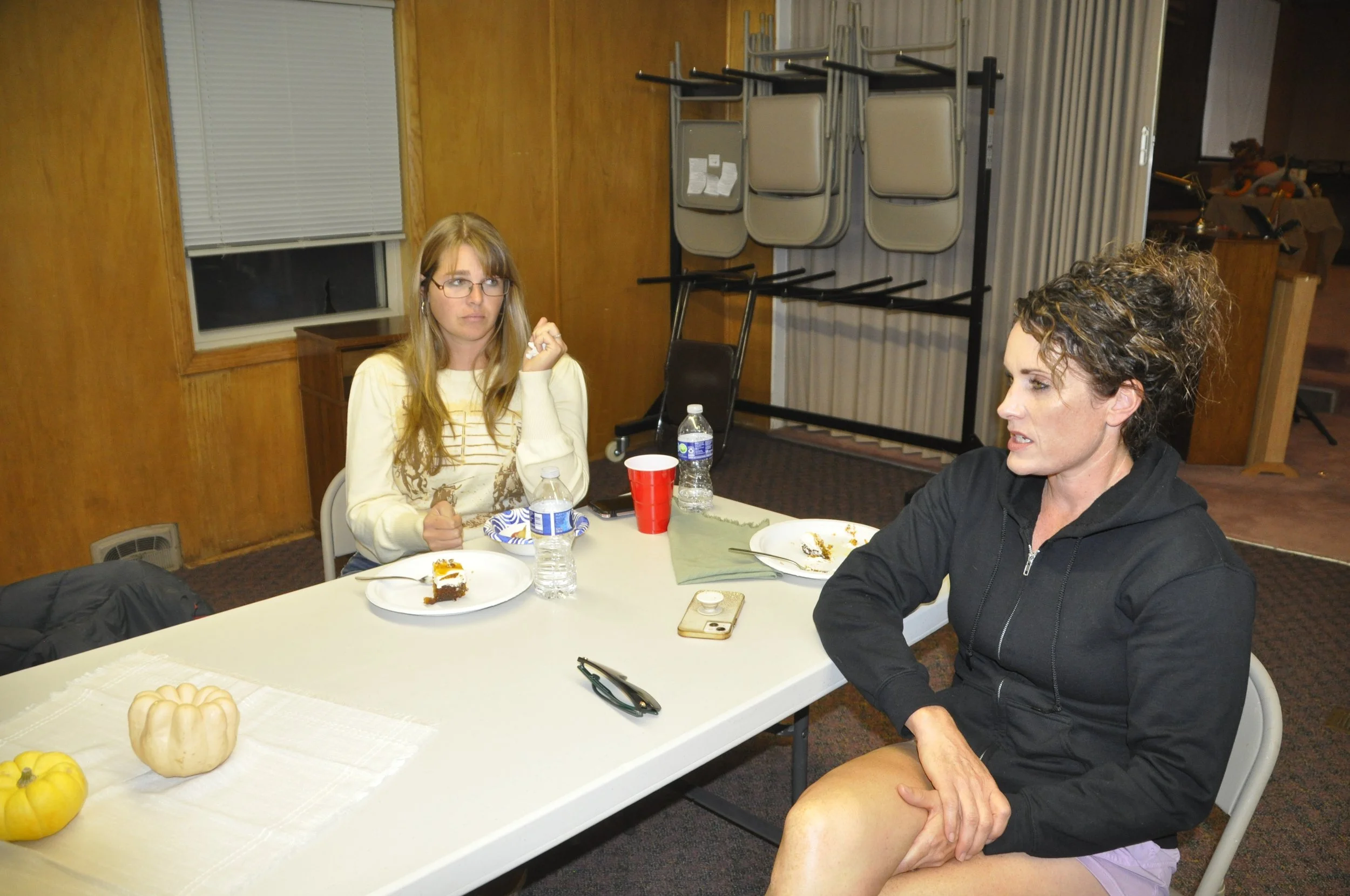 Two women sitting at a table having a conversation. The table has plates with cake, water bottles, a red cup, and a white pumpkin decoration. The room has wood-paneled walls, stacked chairs, and a window with blinds.