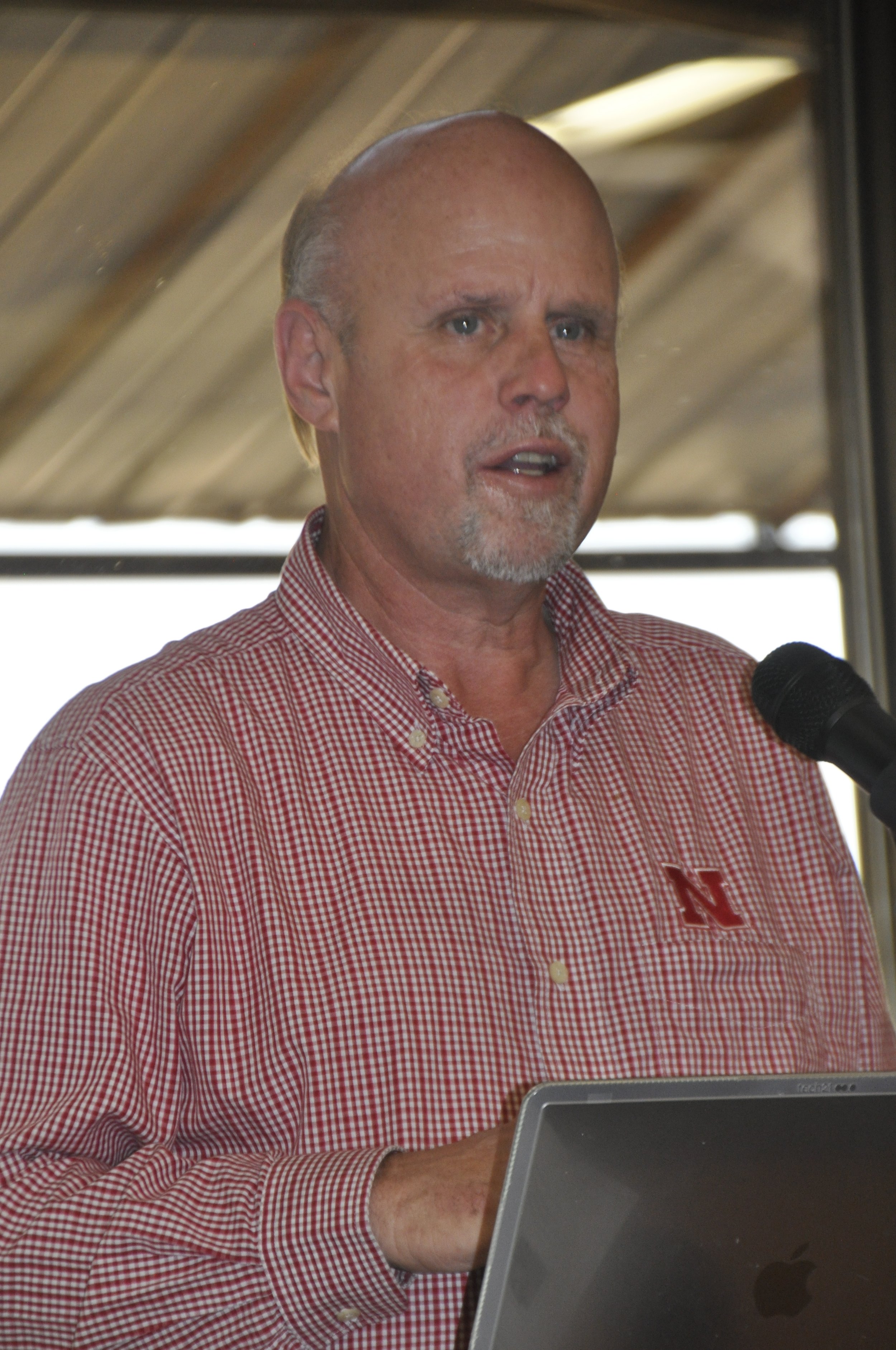 A middle-aged man with a shaved head and facial hair speaking into a microphone during a presentation, wearing a red and white checkered shirt with an 'N' logo on it, in front of a window with a wooden roof structure visible outside.