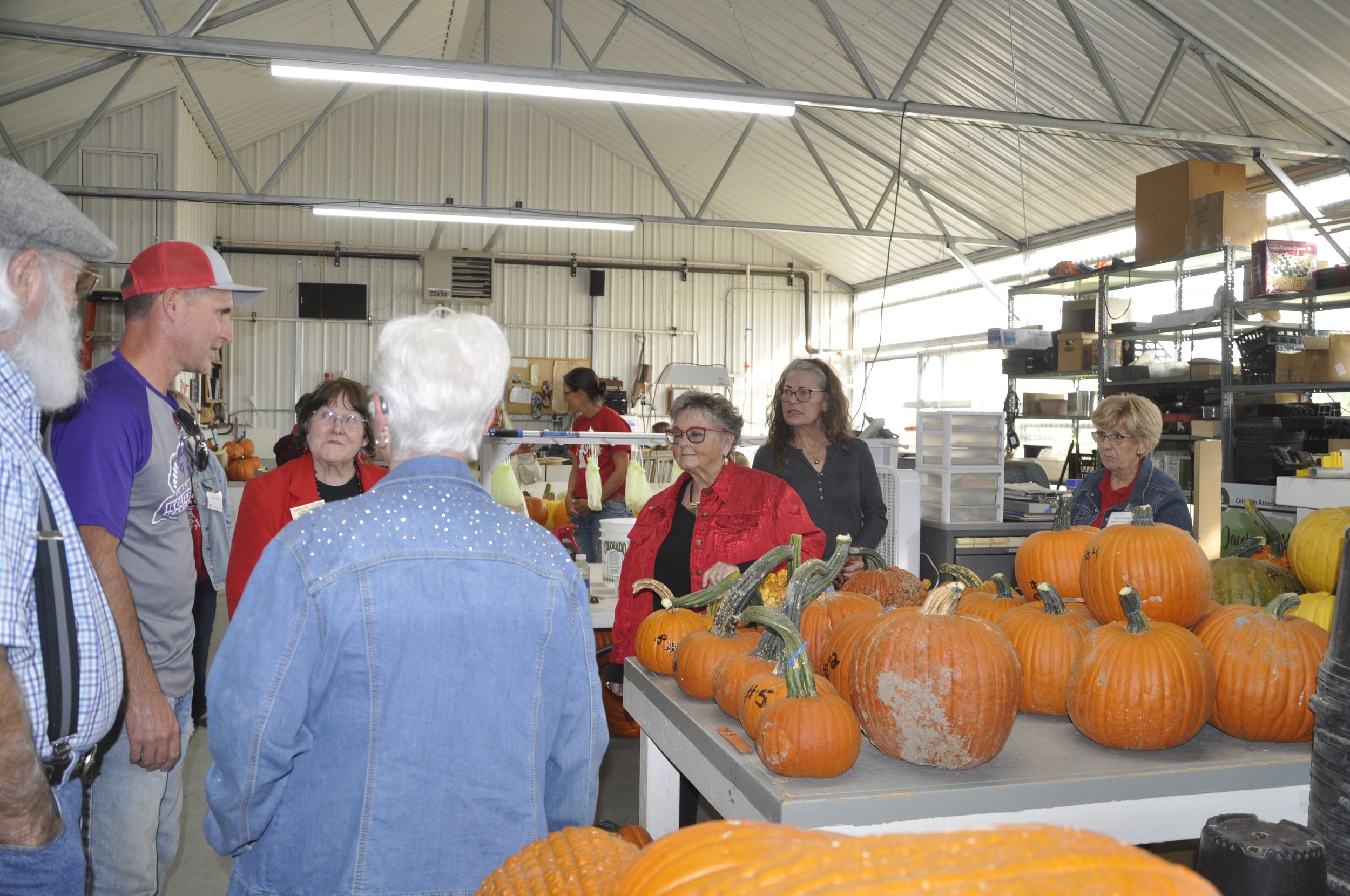 People gathered in a warehouse-like space with a high metal ceiling, around a table of orange pumpkins for sale during a fall event.