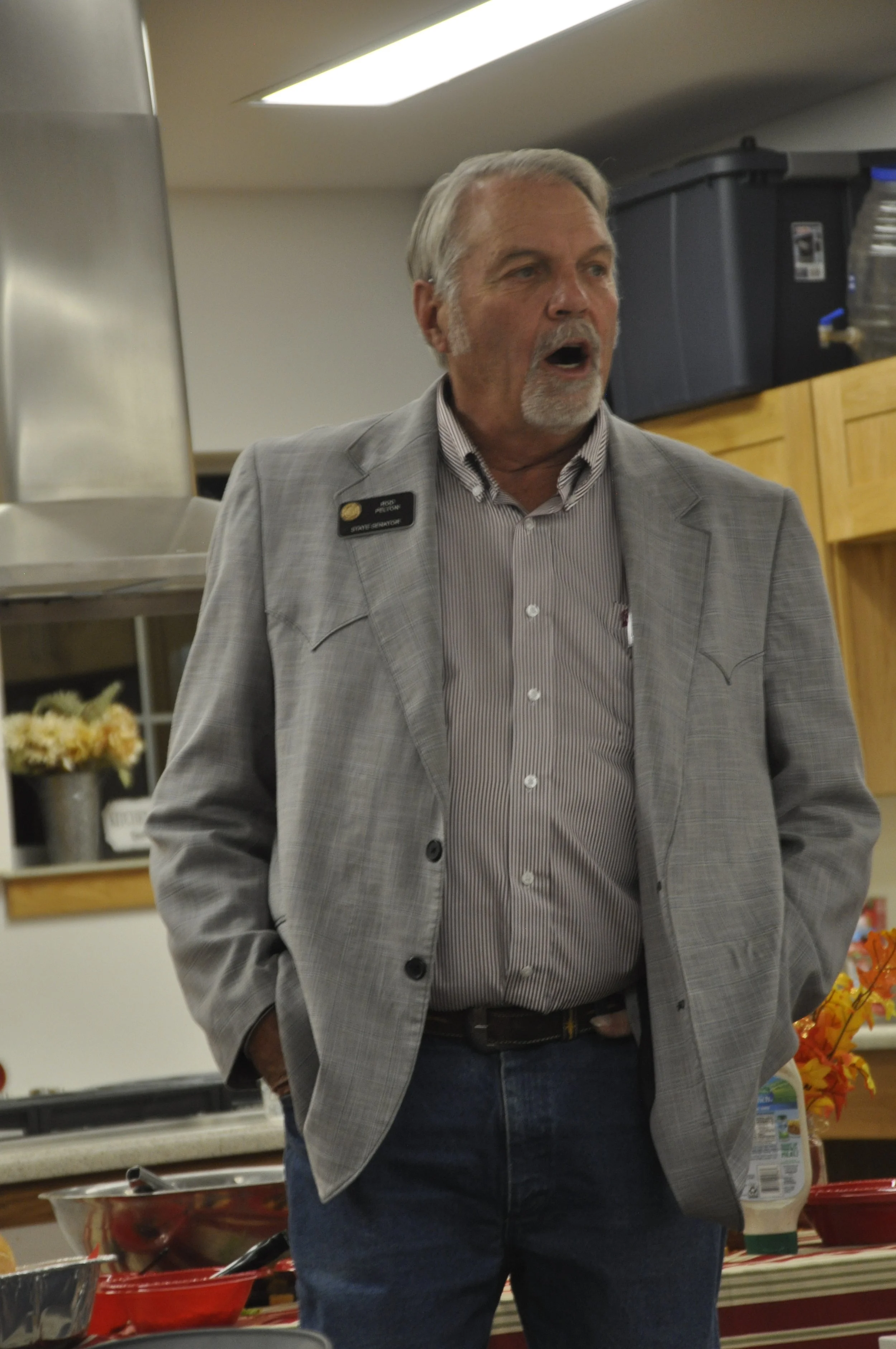 A man with gray hair and a beard, wearing a gray plaid blazer and striped shirt, is speaking in a kitchen or food service area. He has a name tag, and there are food containers and flowers visible in the background.
