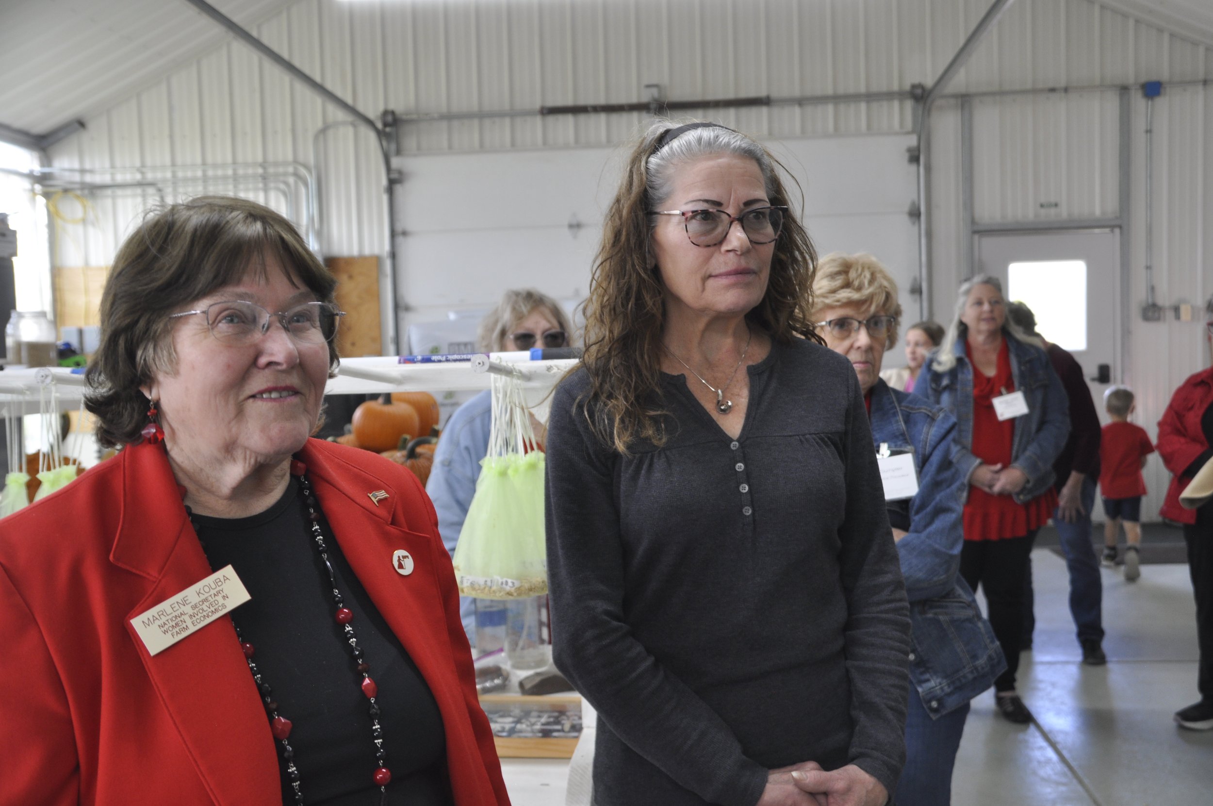 Several women standing in a large, well-lit indoor space with high ceilings. The woman closest to the camera on the left is wearing glasses, a red jacket, and a name badge. The woman next to her has long, curly hair, glasses, and is dressed in dark c