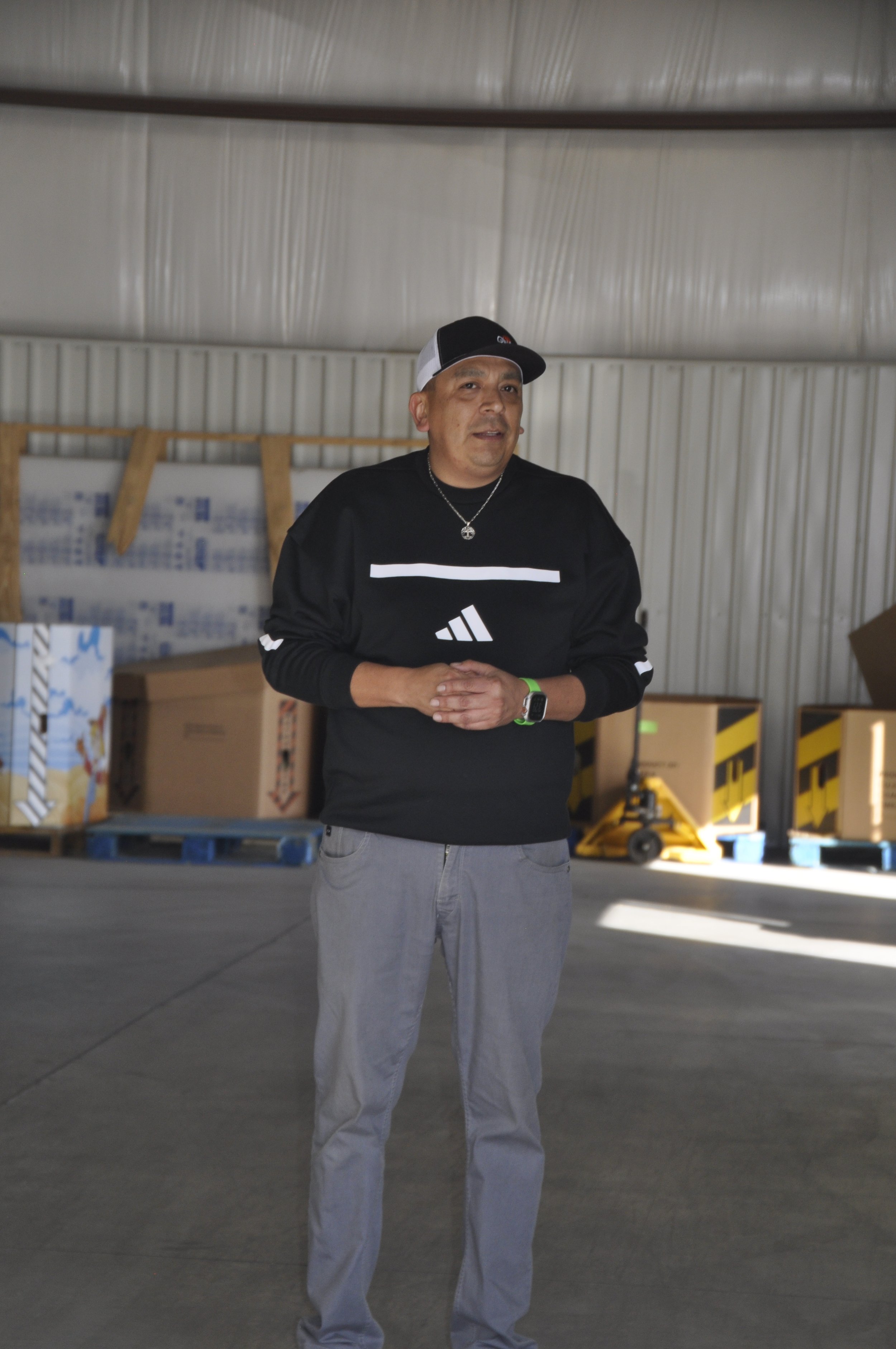A man standing inside a warehouse with a gray industrial ceiling, cardboard boxes, a yellow pallet jack, and black and yellow striped barriers.