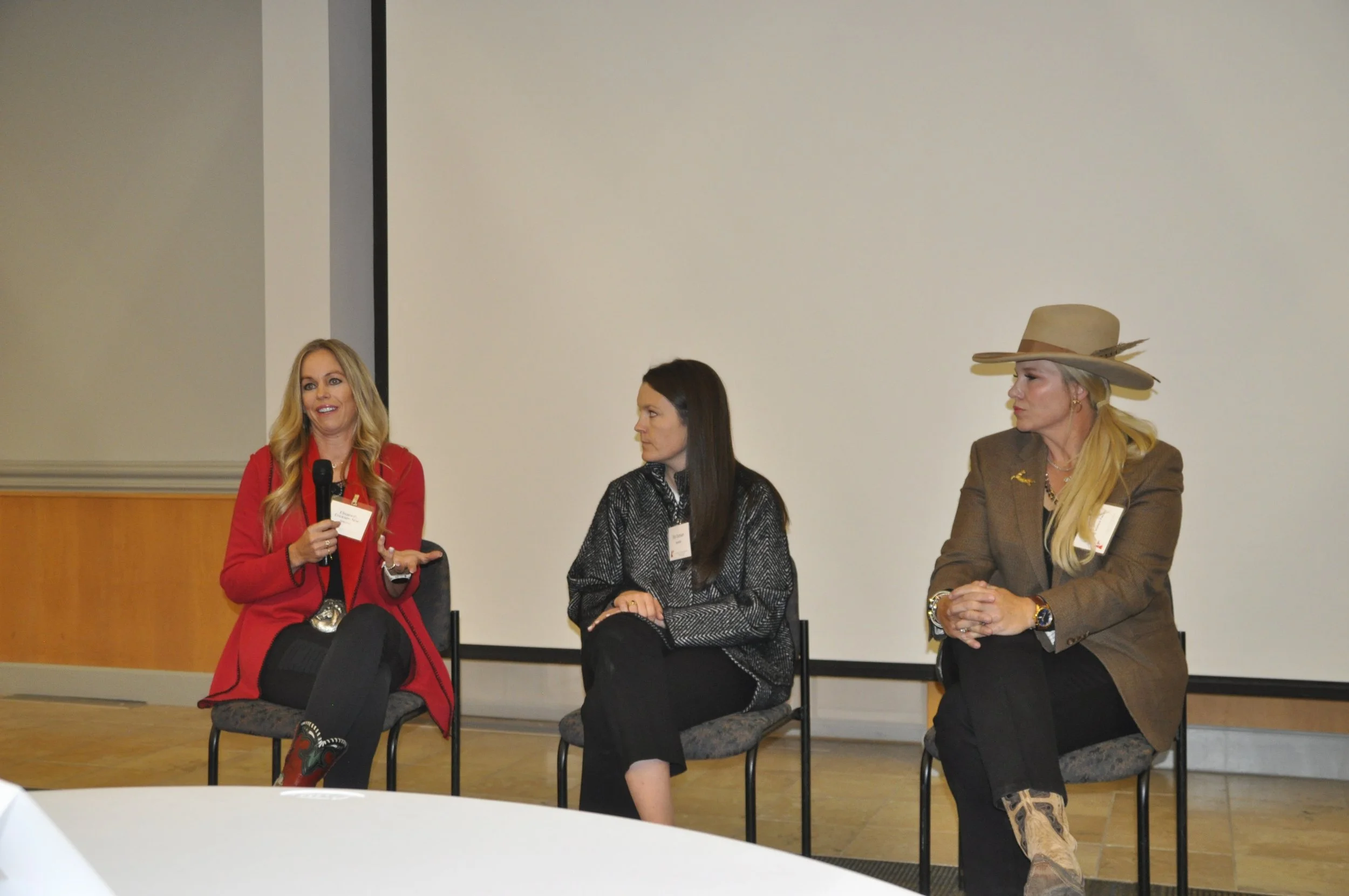 Four women seated on chairs participating in a panel discussion in a conference room. One woman, wearing a red blazer and black pants, is speaking into a microphone, while the other three women are listening. The woman on the far right is wearing a l