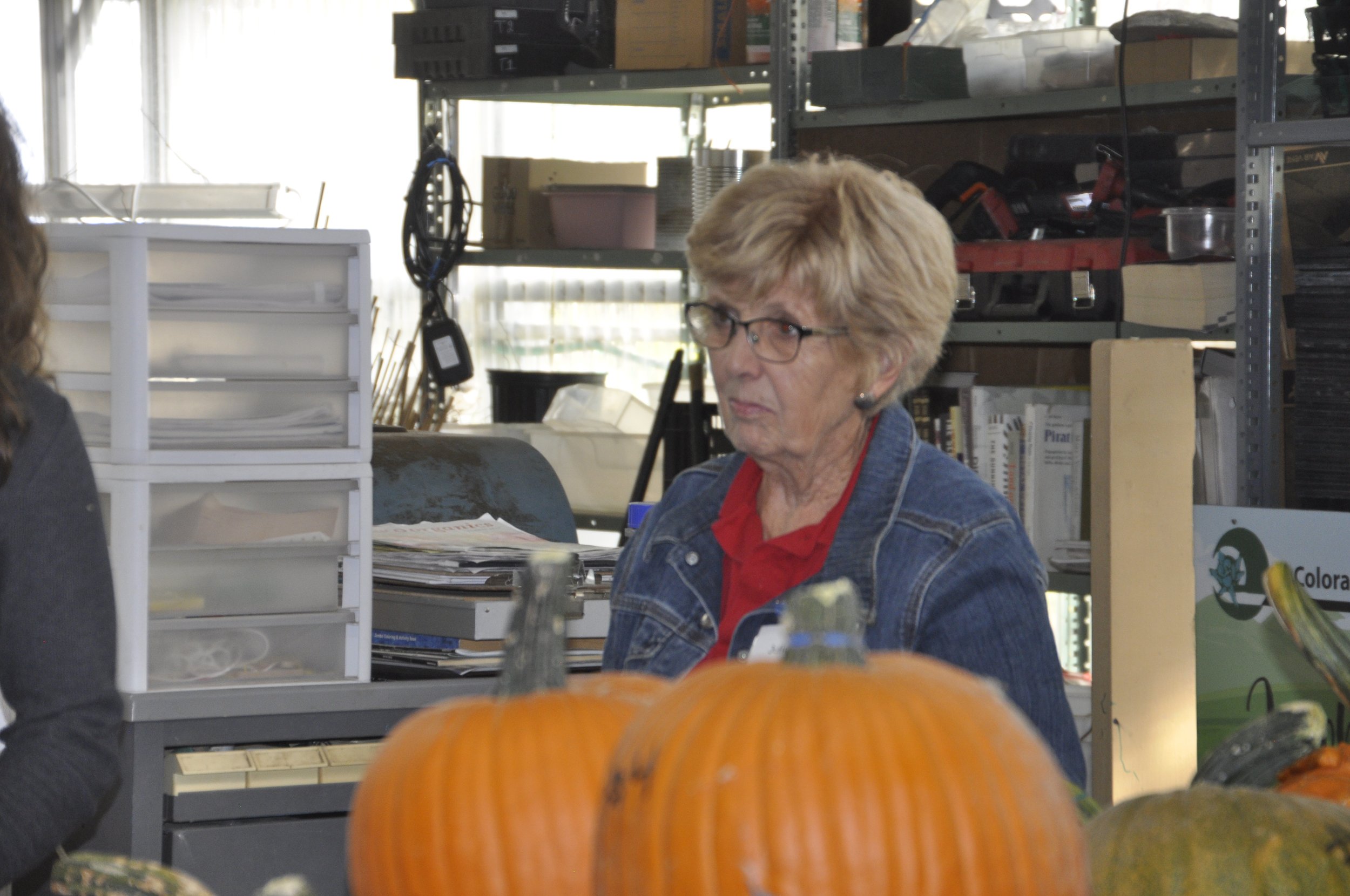 A woman with short blonde hair and glasses, wearing a denim jacket and red shirt, sitting at a desk in a workshop or office filled with shelves and supplies, with pumpkins in the foreground.