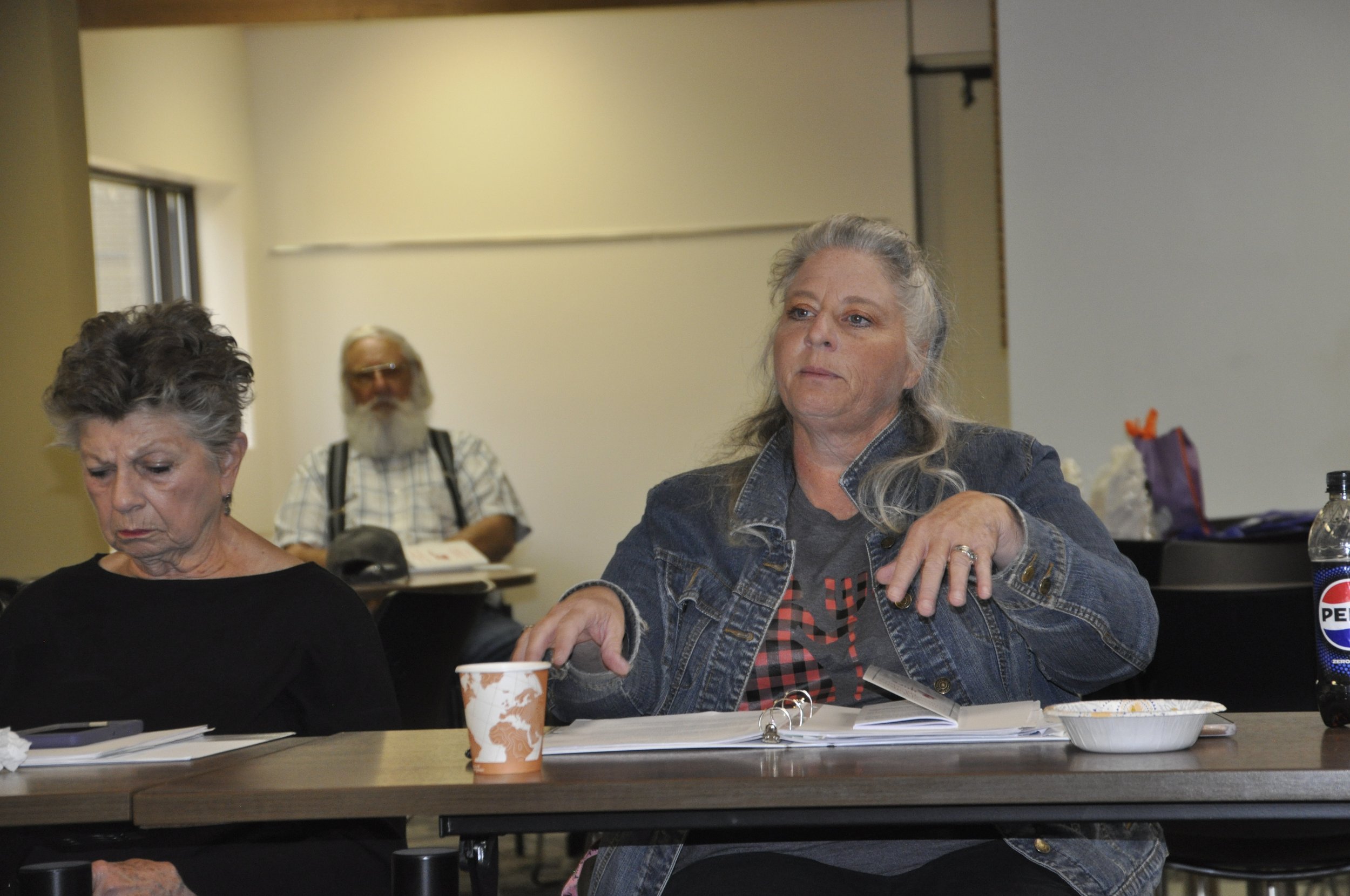 Three older adults seated at a table in a room, with papers, notebooks, a cup, and a water bottle in front of them. The woman in the foreground has gray hair, is wearing a denim jacket, and is reaching for something on the table. The woman on the lef