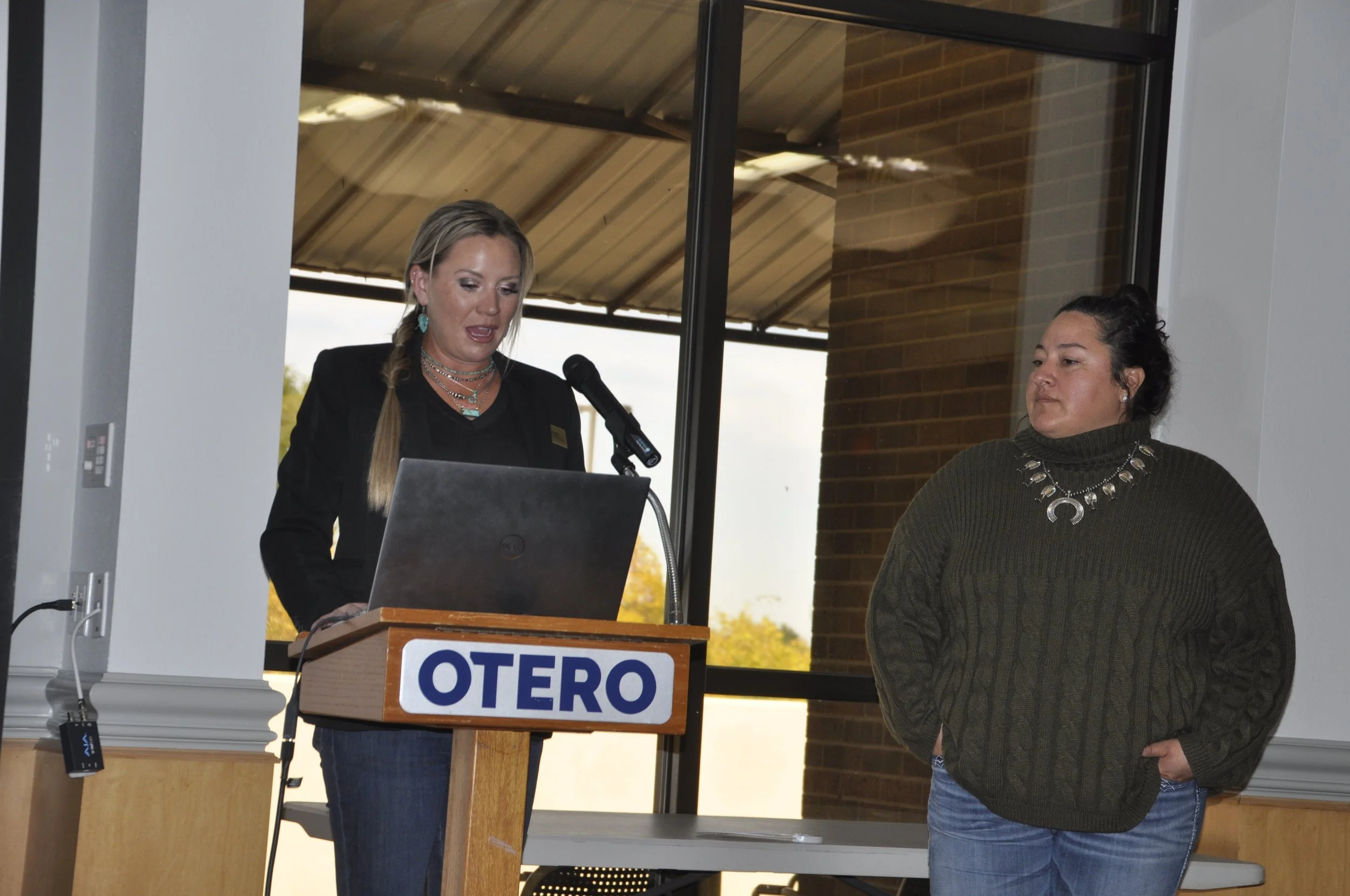 Two women in a room with large windows, one standing at a podium with a laptop and the other standing beside her. The podium has a sign that says 'OTERO'. The woman at the podium has long blonde hair, is wearing a black blazer, and appears to be spea