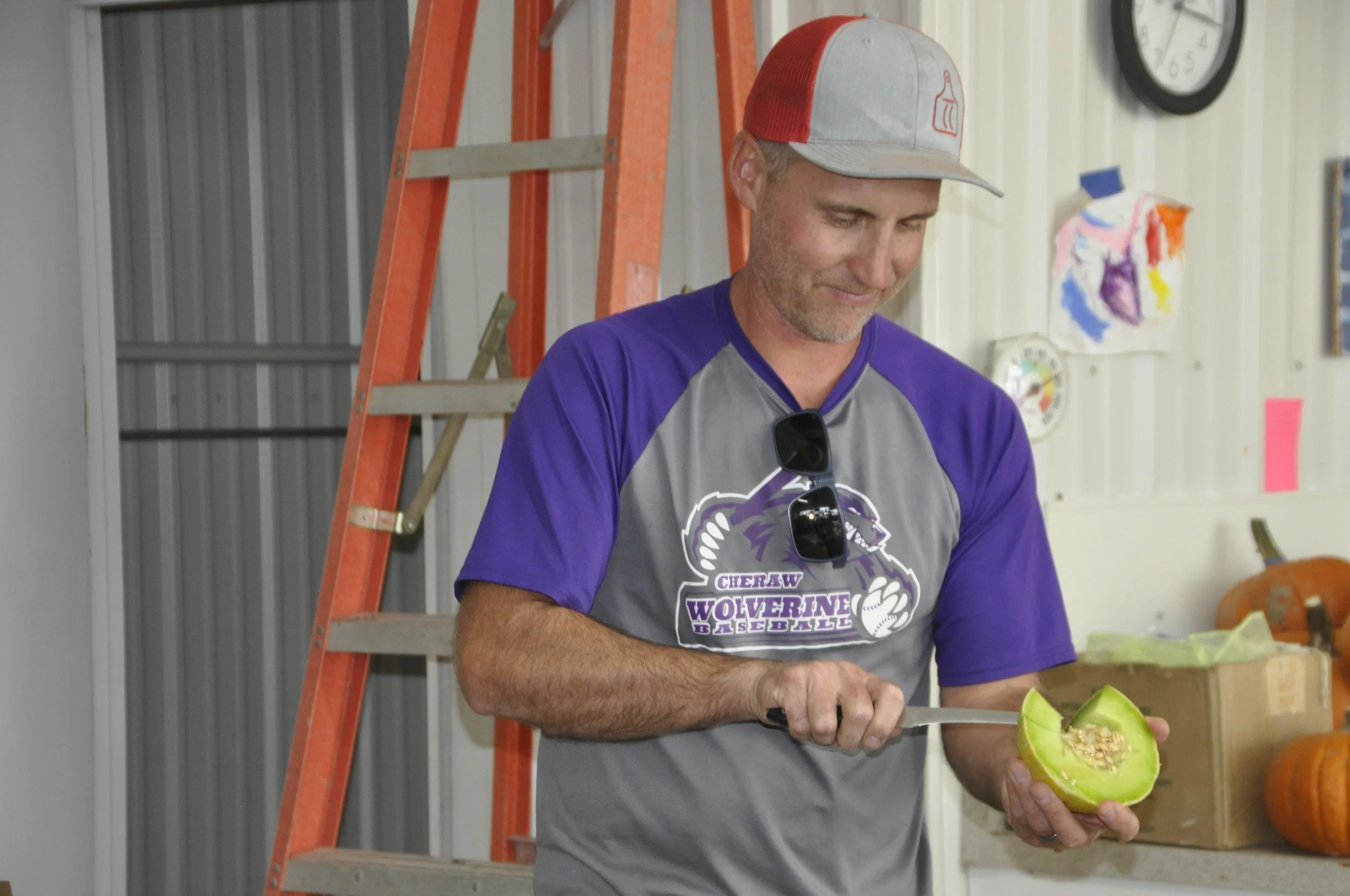 A man is cutting a green melon with a knife in a room decorated with children's artwork and pumpkins.