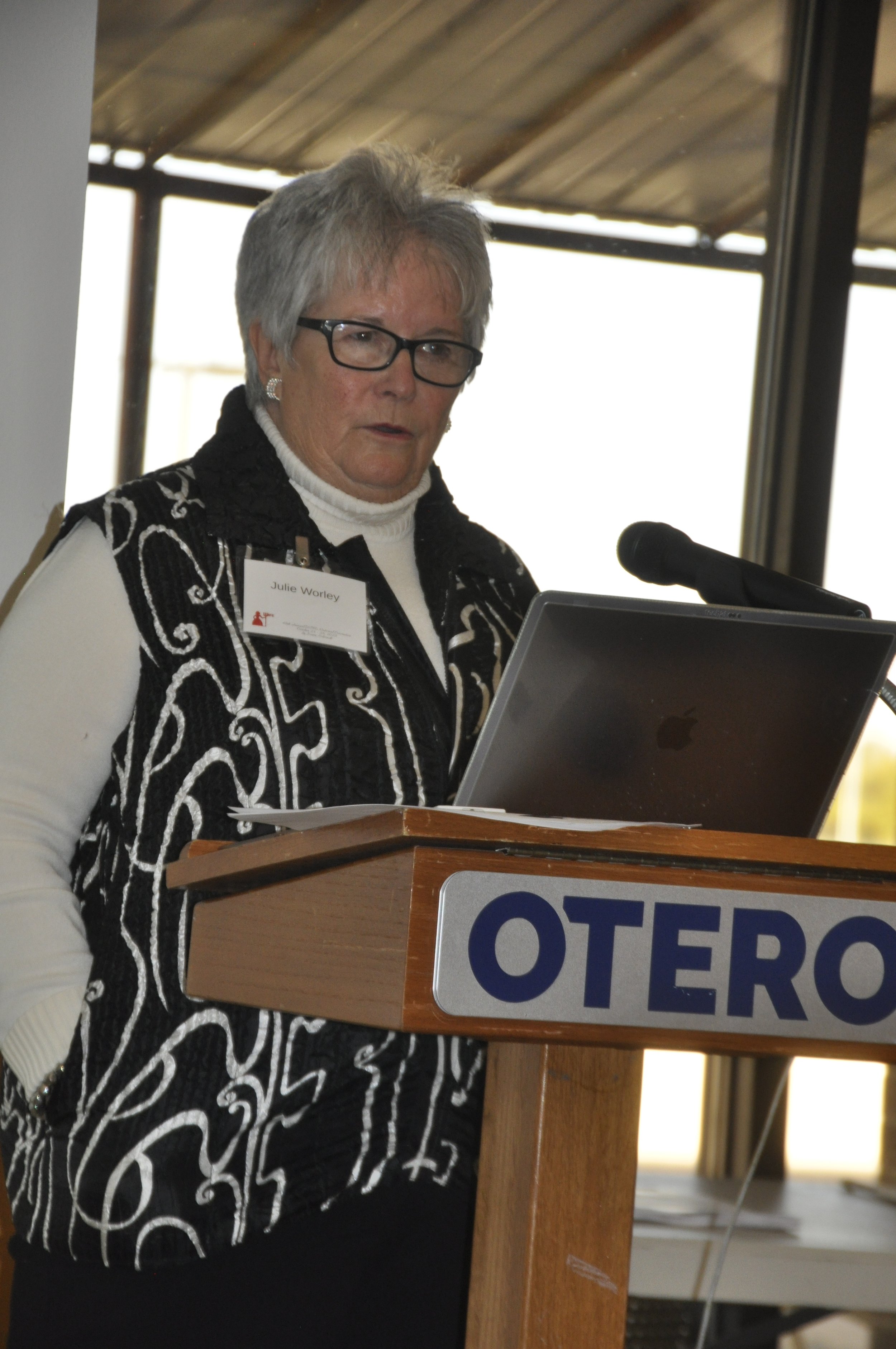 Woman with gray hair and glasses speaking at a podium labeled "OTERO."
