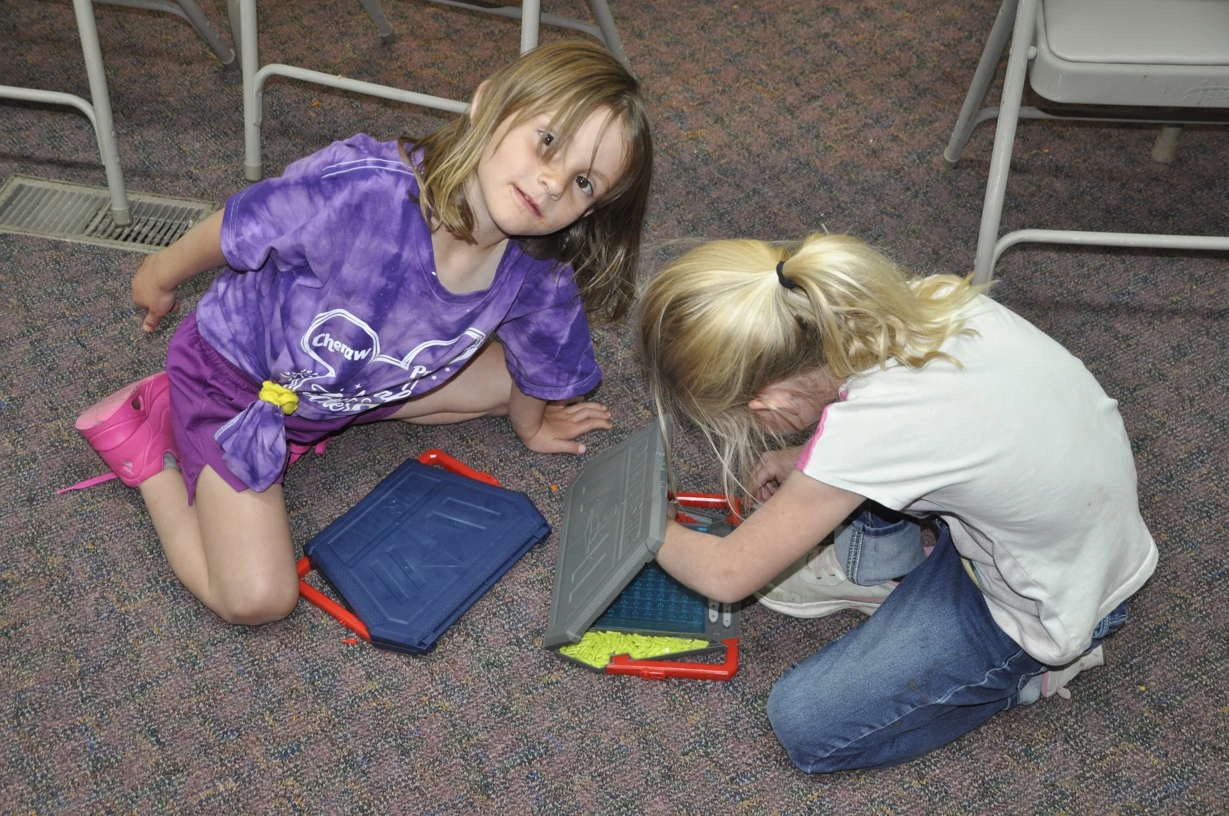 Two young girls are kneeling on a school carpeted floor, working together with a plastic toolbox containing yellow connectors and construction tools, surrounded by school chairs.
