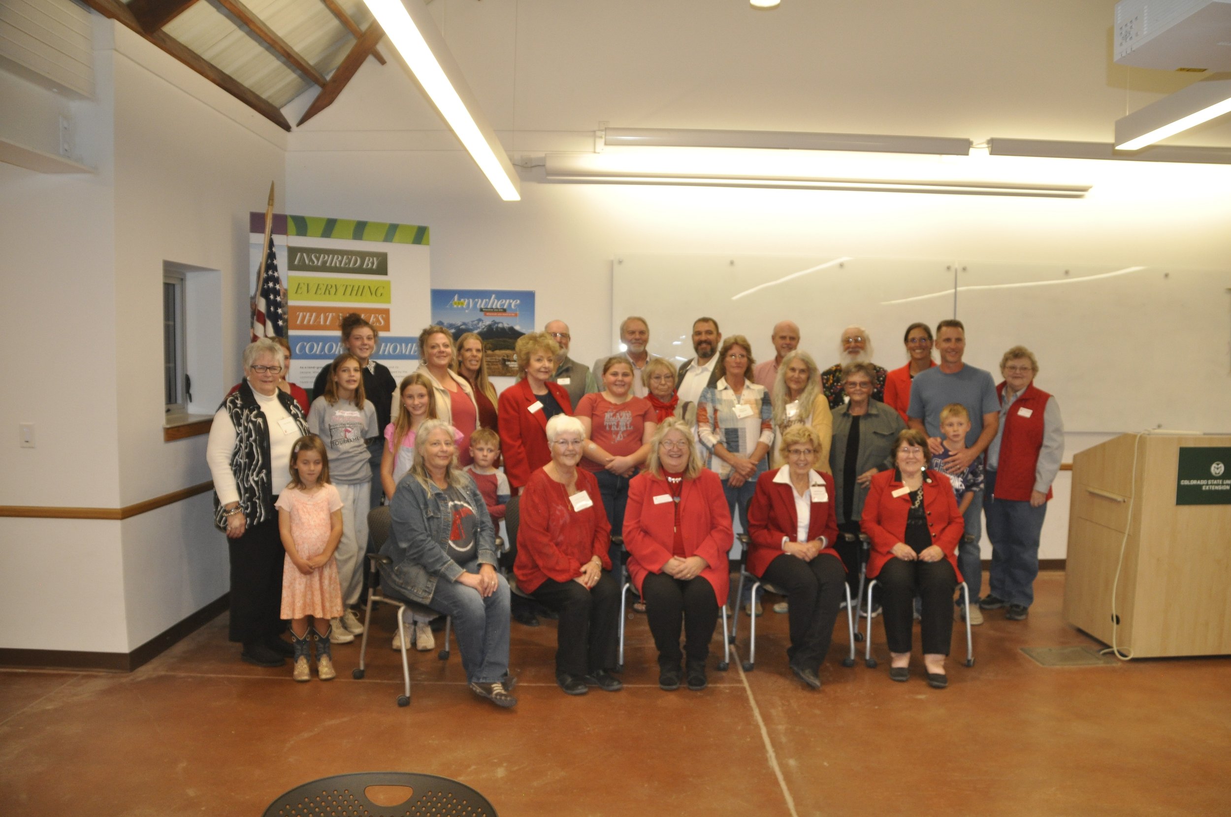 A group of people, mostly women and children, gathered in a room for a photo. Some are dressed in red and others in casual attire. There are banners in the background, including one that reads 'Inspired by Everything that makes Colorado home.'