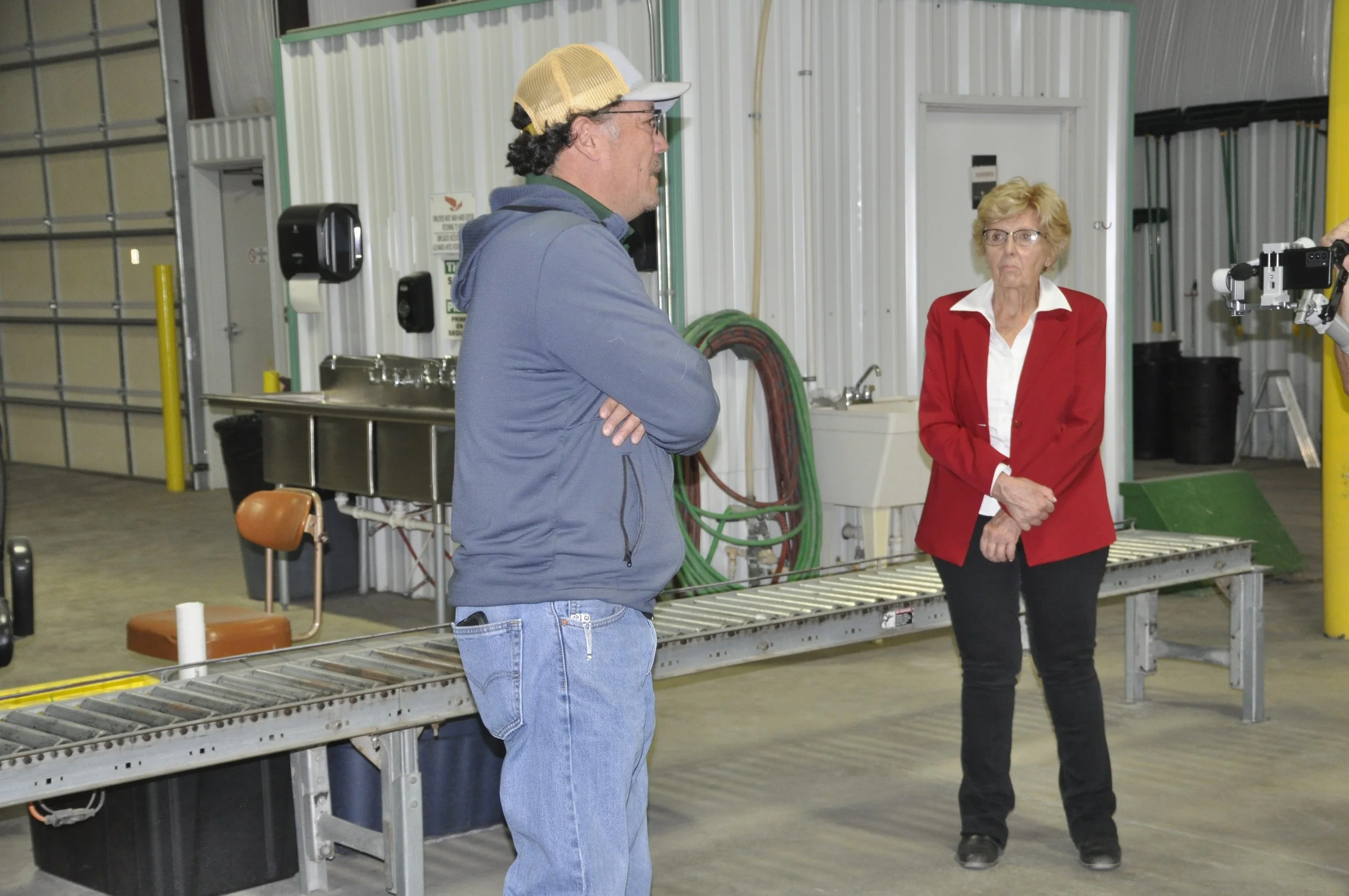 Two people, a man with a beige cap and a woman with glasses, standing and talking in an industrial or warehouse setting with metal walls, green hoses, and equipment nearby.