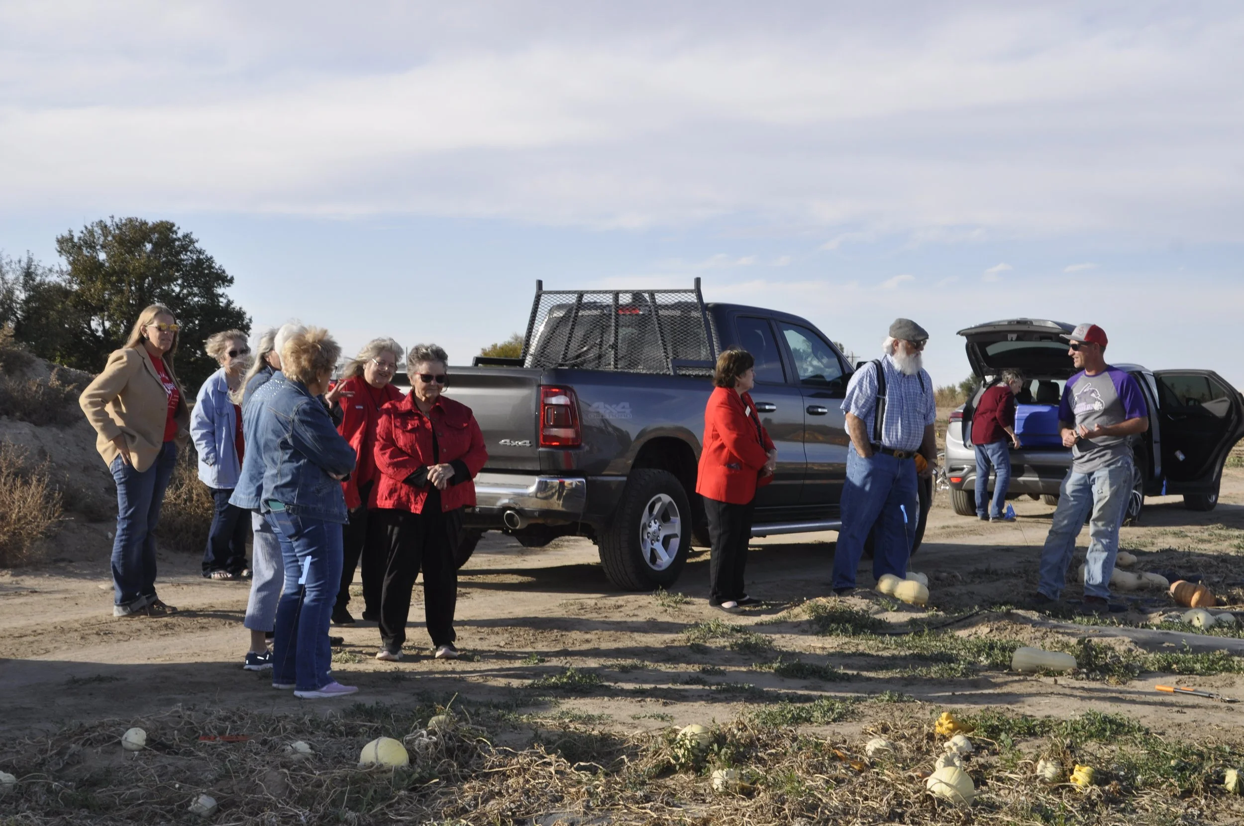 A group of people, mostly women, standing outdoors near parked trucks, with some pumpkins on the ground, under a partly cloudy sky.