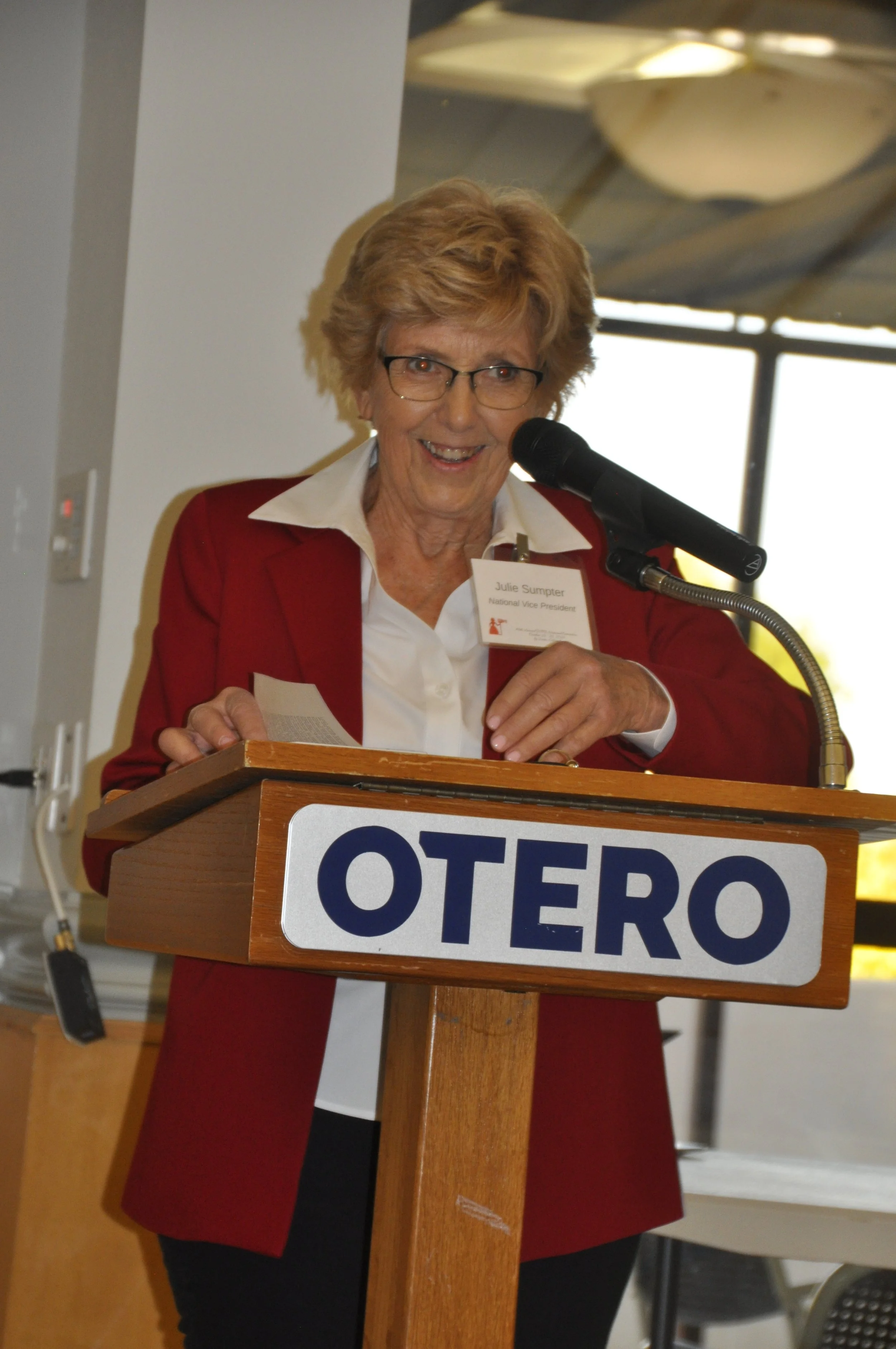 A woman speaking at a podium labeled "OTERO" during a formal event. She is wearing glasses, a white shirt, and a red blazer, and has a name badge that reads 'Julie Sumpter, National Vice President.' She is smiling and speaking into a microphone.