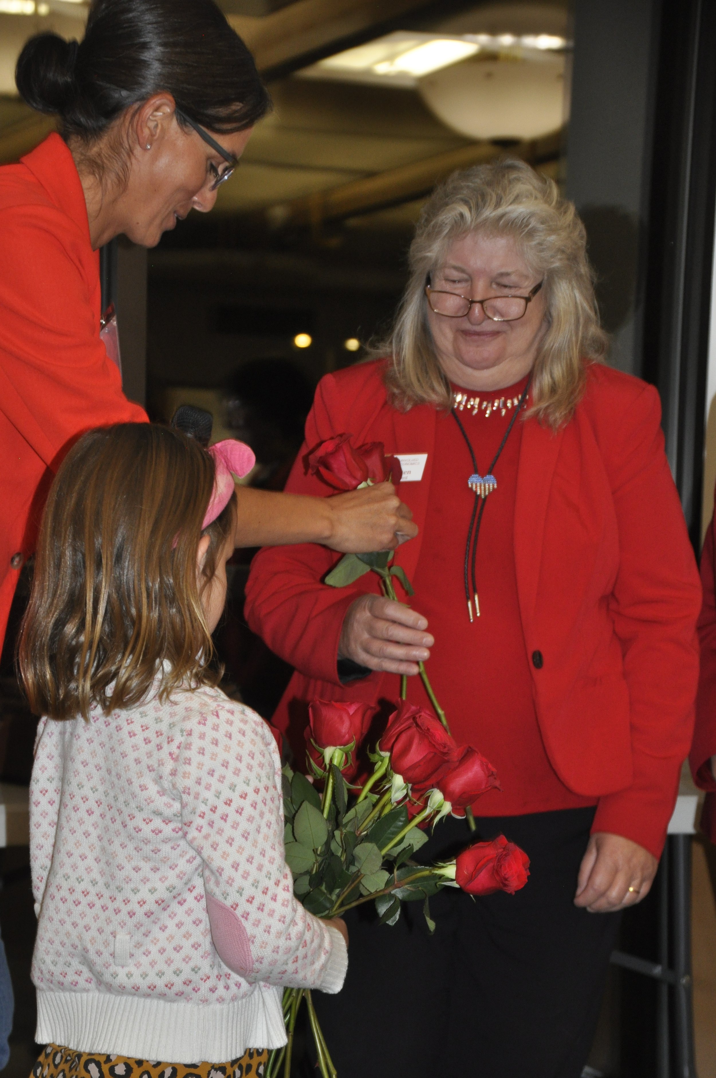 A woman in a red outfit holding a bouquet of red roses, giving a flower to a young girl with long brown hair and a pink bear headband, in an indoor setting with a woman wearing glasses in the background.