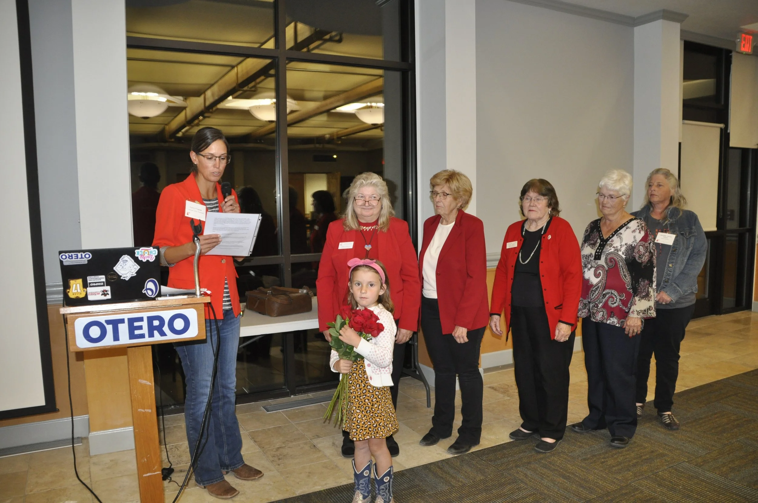 A woman speaking into a microphone at a podium with a sign reading 'OTERO,' surrounded by a group of women and a young girl holding a bouquet of red roses at an indoor event.