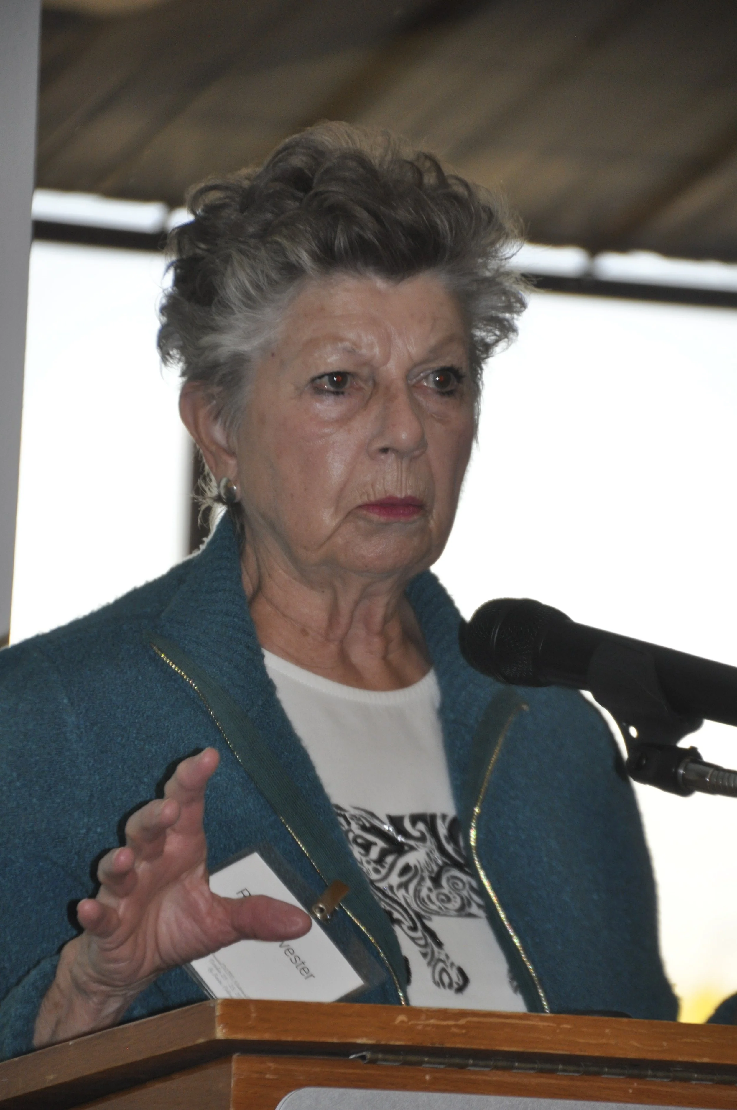 An elderly woman with short gray hair and wearing a teal jacket, speaking into a microphone at a podium, gesturing with her right hand, during a speech or presentation.