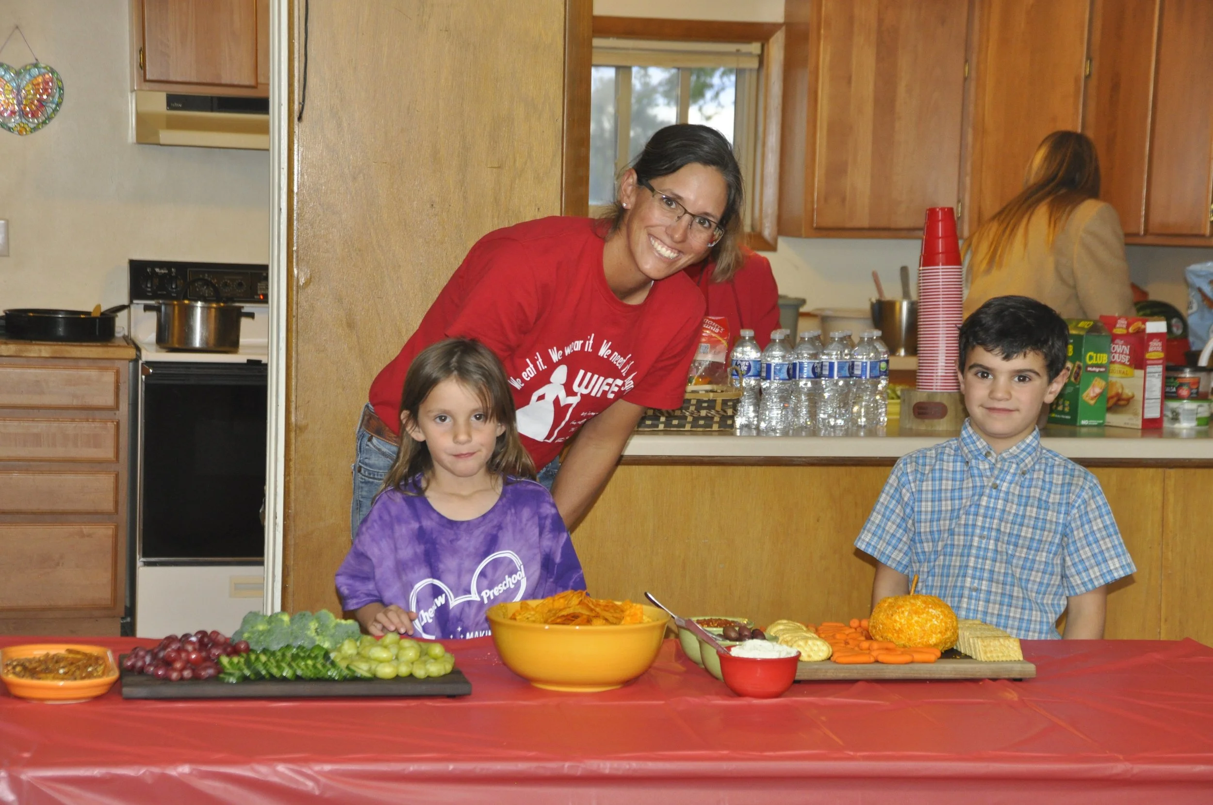 A woman and two children are at a table with various healthy snacks and fruits, in a kitchen kitchen with wooden cabinets and a window. The woman is smiling, wearing glasses and a red shirt, while the girl in a purple shirt and the boy in a plaid shi