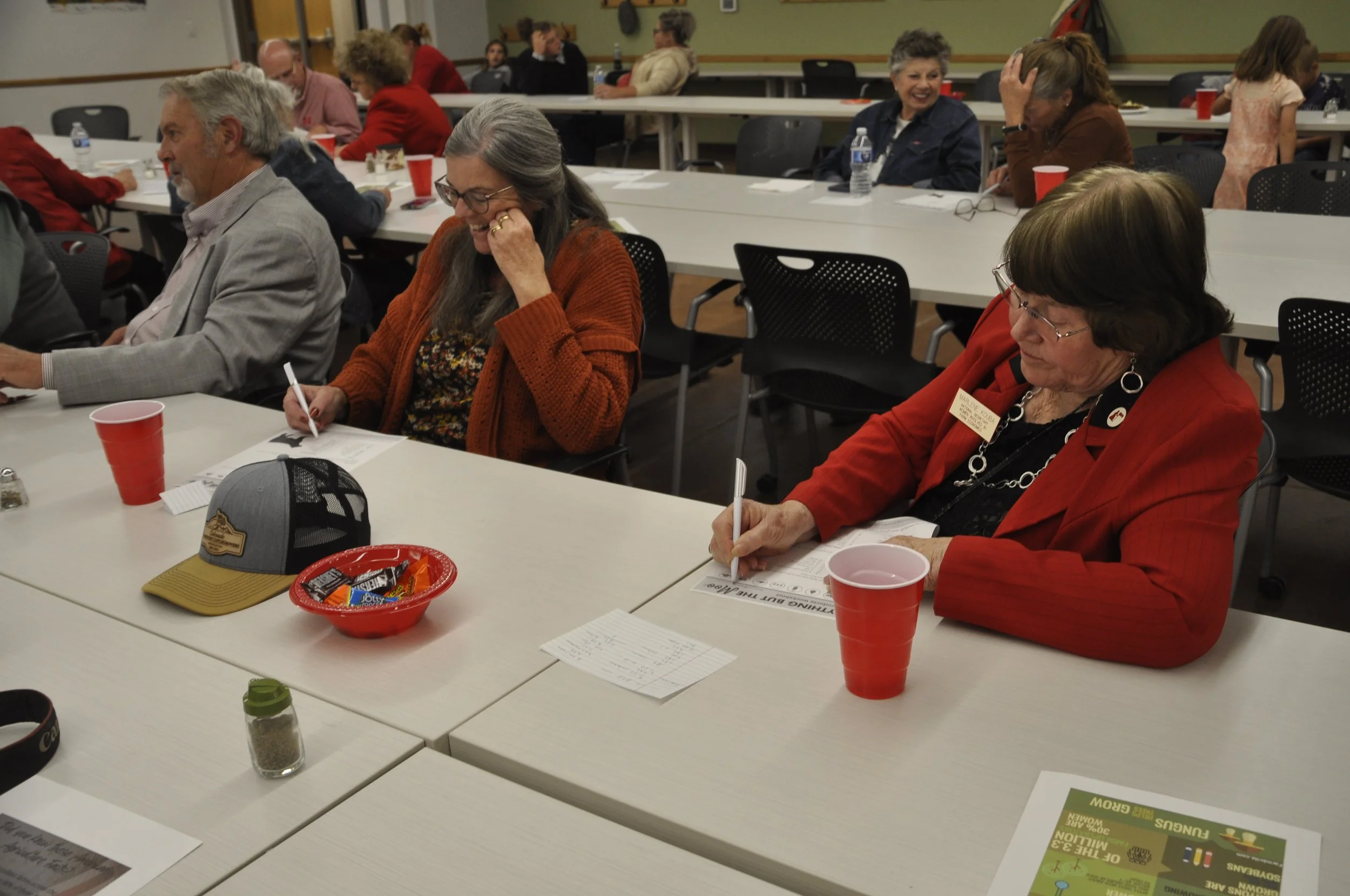 People attending a meeting or workshop in a room, seated at white tables with papers, pens, and red cups, some writing or reviewing documents, and engaging with each other.