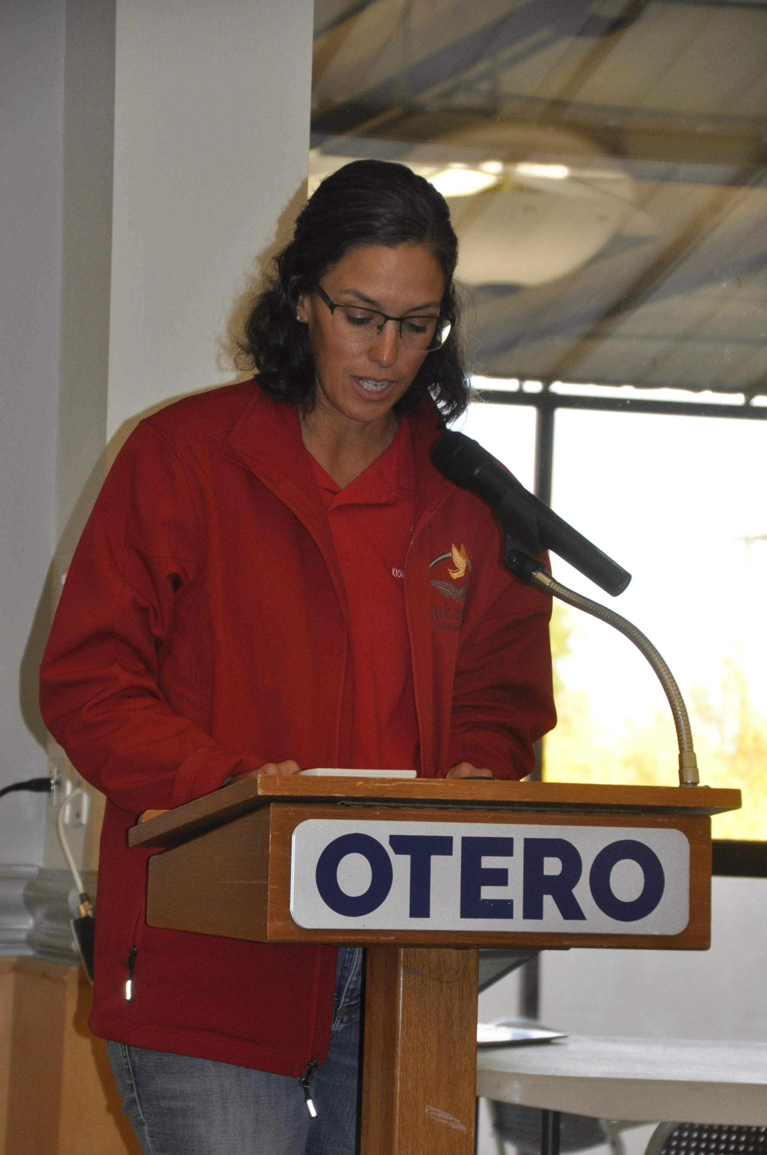 A woman with glasses and dark hair, wearing a red jacket, standing at a podium with a sign that says OTERO, speaking into a microphone.