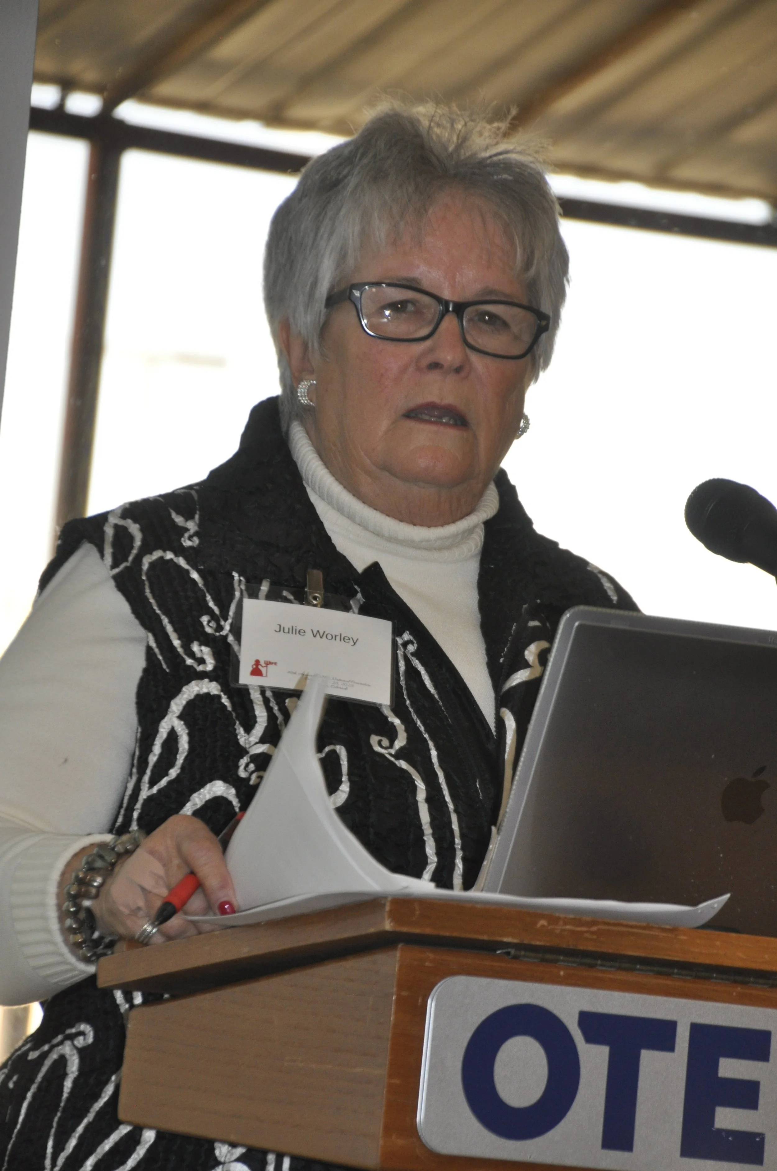 A woman with short gray hair, wearing black glasses, a black and white patterned jacket, and a white turtleneck, stands at a podium, speaking into a microphone and holding papers. She is wearing earrings and a bracelet, and is labeled as Julie Worley