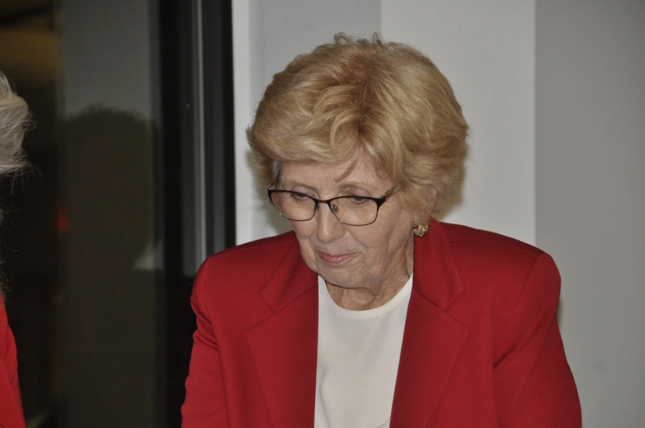 An elderly woman with short, curly, reddish hair and glasses wears a red blazer and white shirt, looking down, indoors with a plain wall background.