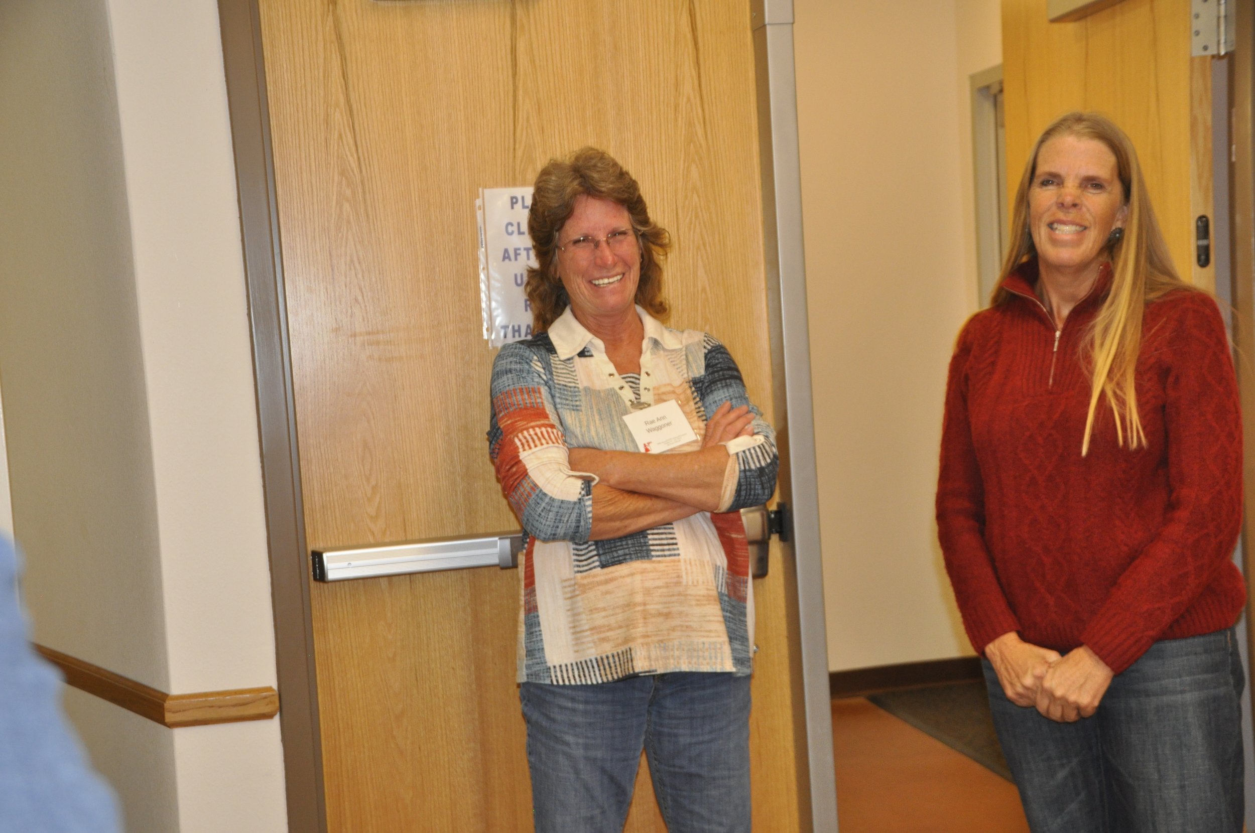 Two women standing in front of a wooden door, smiling, at an indoor event. The woman on the left has curly hair, glasses, and is wearing a patterned cardigan. The woman on the right has long straight hair and is wearing a red sweater.