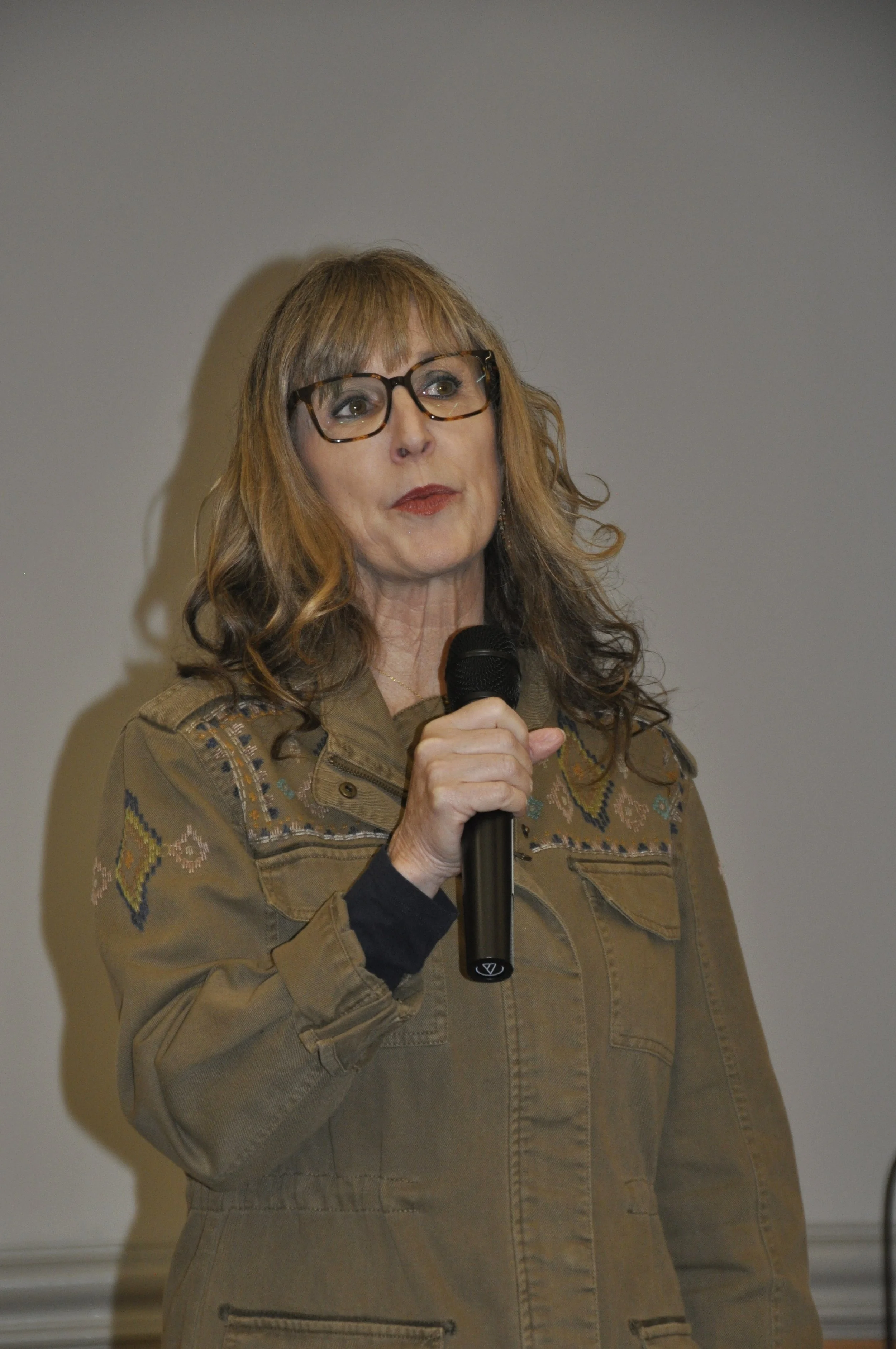 A woman with long, wavy, light brown hair and glasses speaking into a microphone in a beige jacket with embroidery.