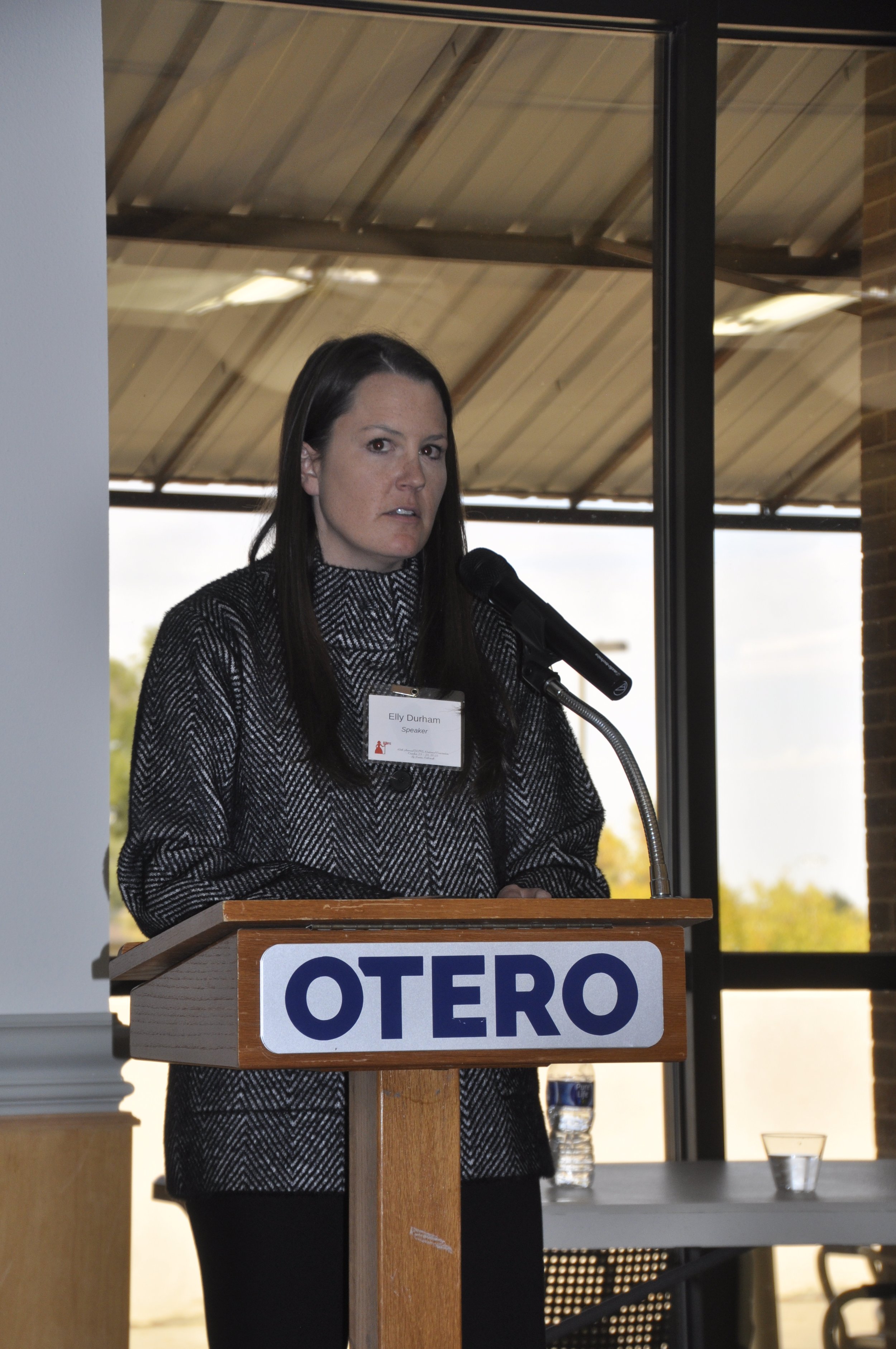 A woman standing at a podium, delivering a speech, with a window behind her showing an outdoor scene and a roof structure overhead.