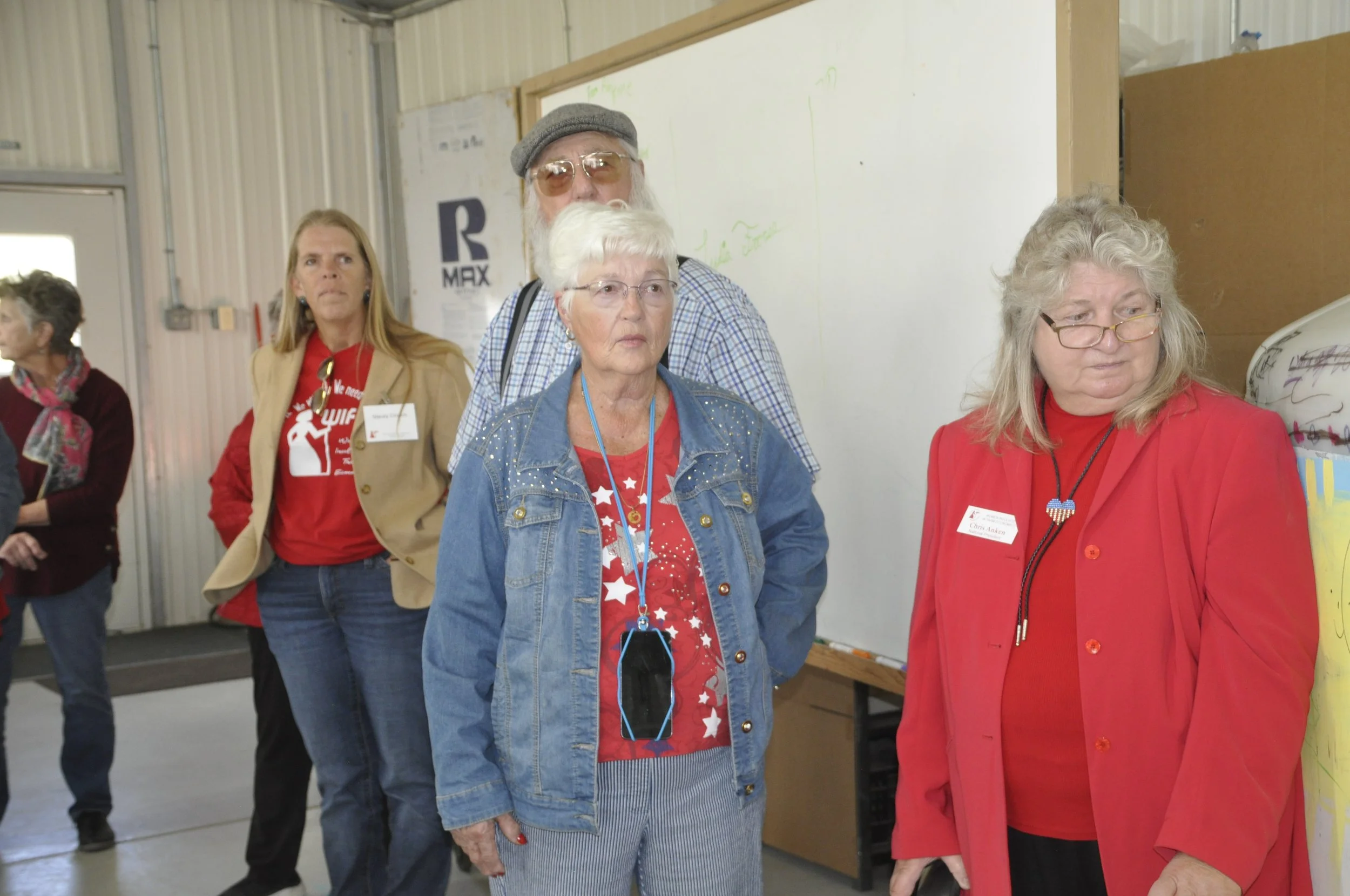 Group of women standing indoors, some wearing red shirts with patriotic designs, and one in a tan jacket. They are looking towards the right, in front of a whiteboard and a wall with various papers and notes.