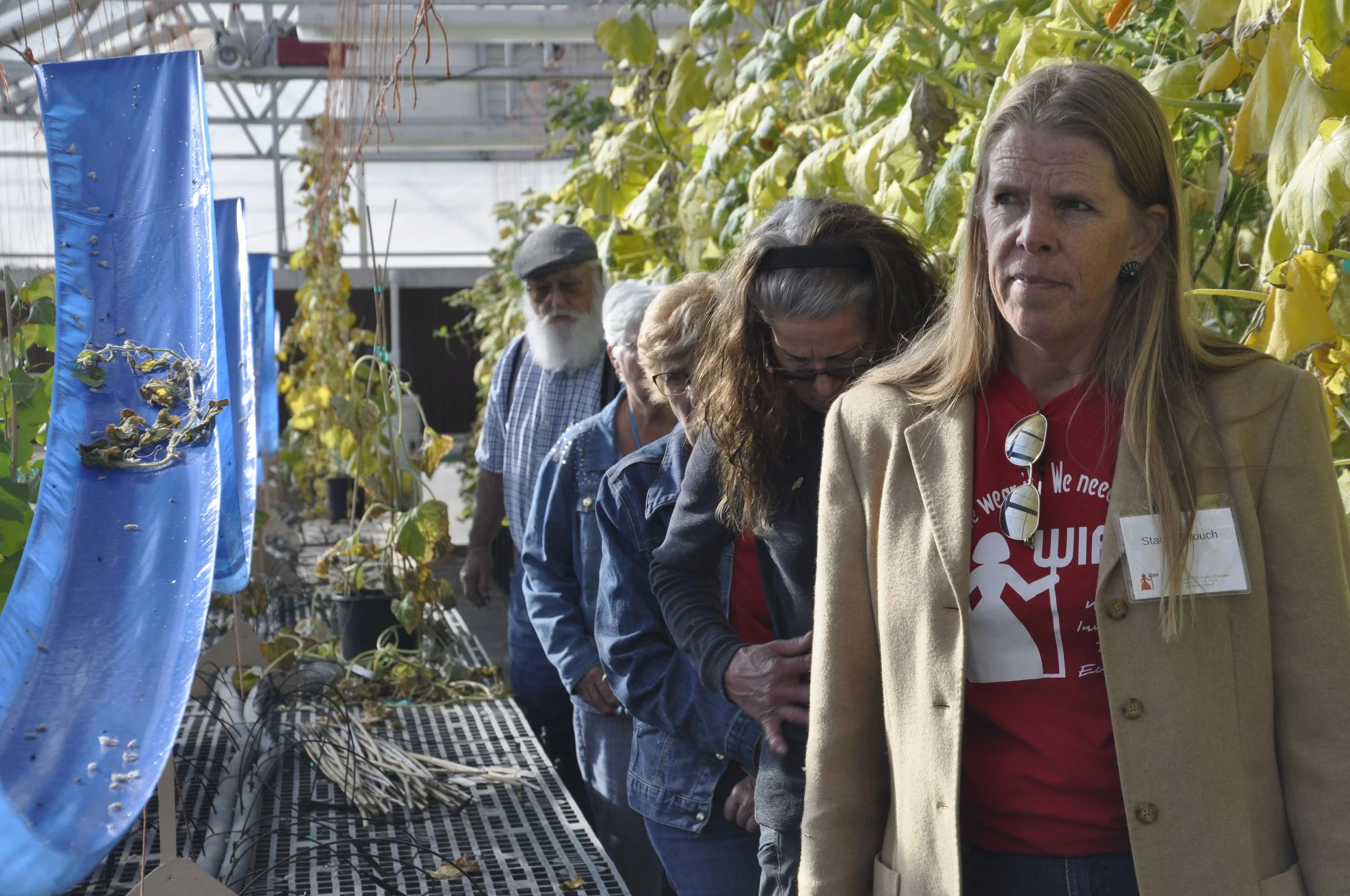 People standing in a line inside a greenhouse or plant nursery with large green foliage and blue side panels.