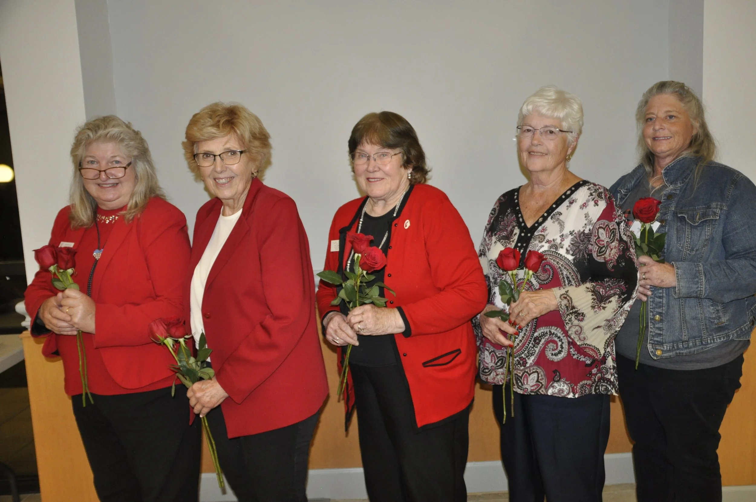Five women standing in a line, each holding a red rose, smiling, and looking at the camera against a neutral background.
