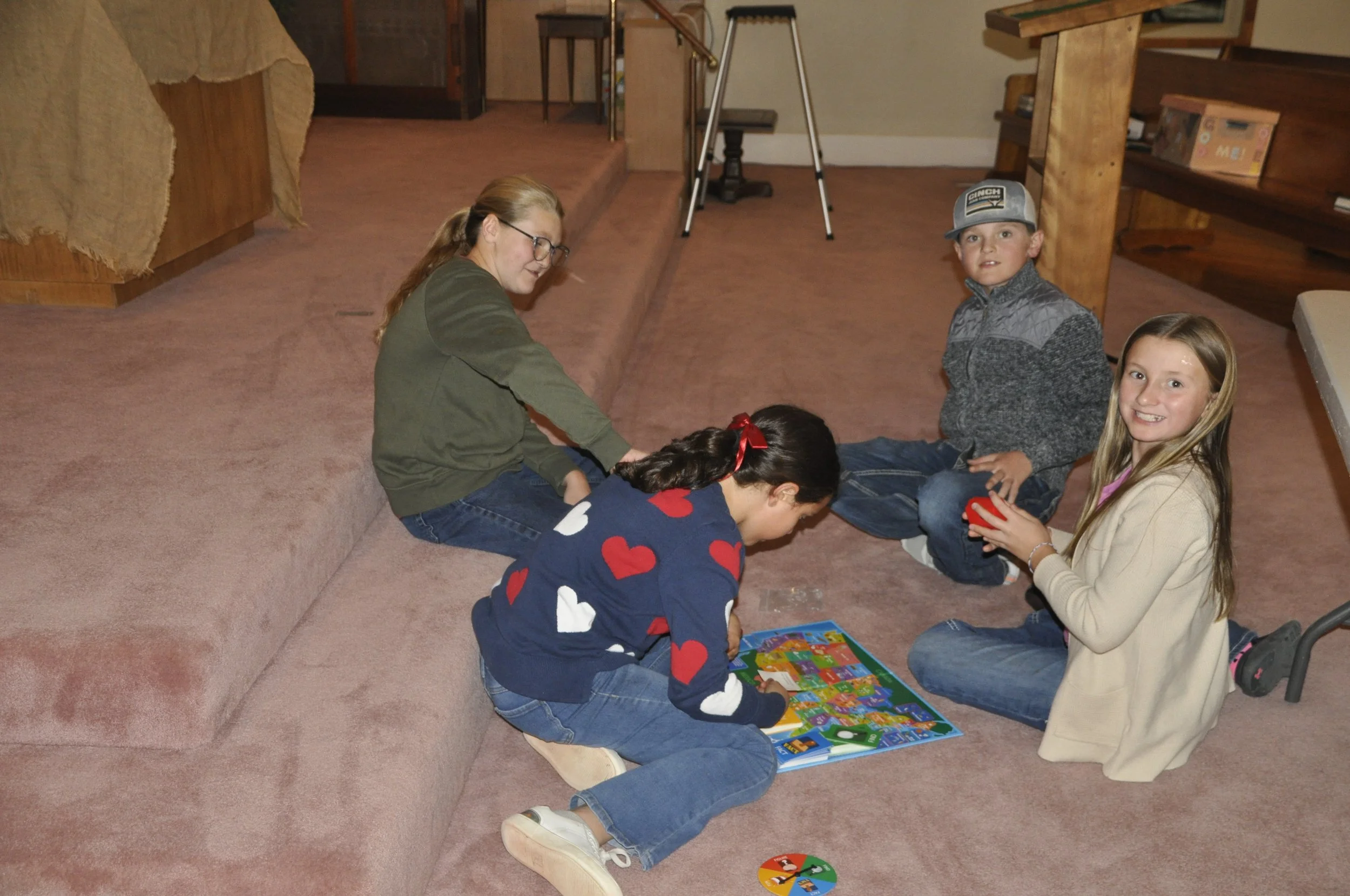 A group of five kids sitting on the carpeted floor of a room, engaged in playing board games. One girl with long hair and glasses is smiling, while another girl with long blonde hair holds a game piece. Two boys are sitting nearby, one wearing a base