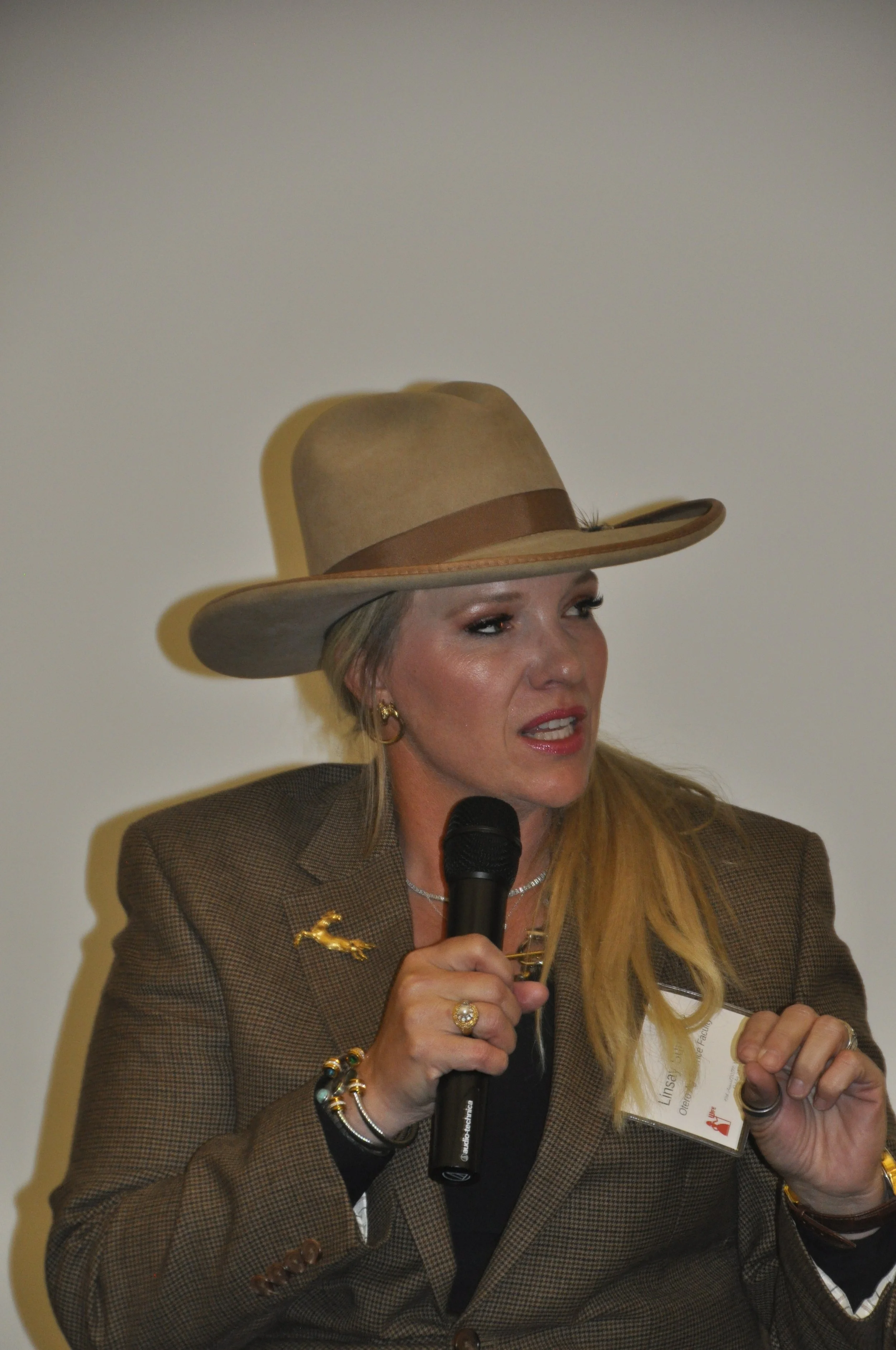 A woman with blonde hair, wearing a brown blazer, holding a microphone and a name badge, speaking at an event, wearing a tan wide-brimmed hat with a brown band and jewelry, against a plain wall background.