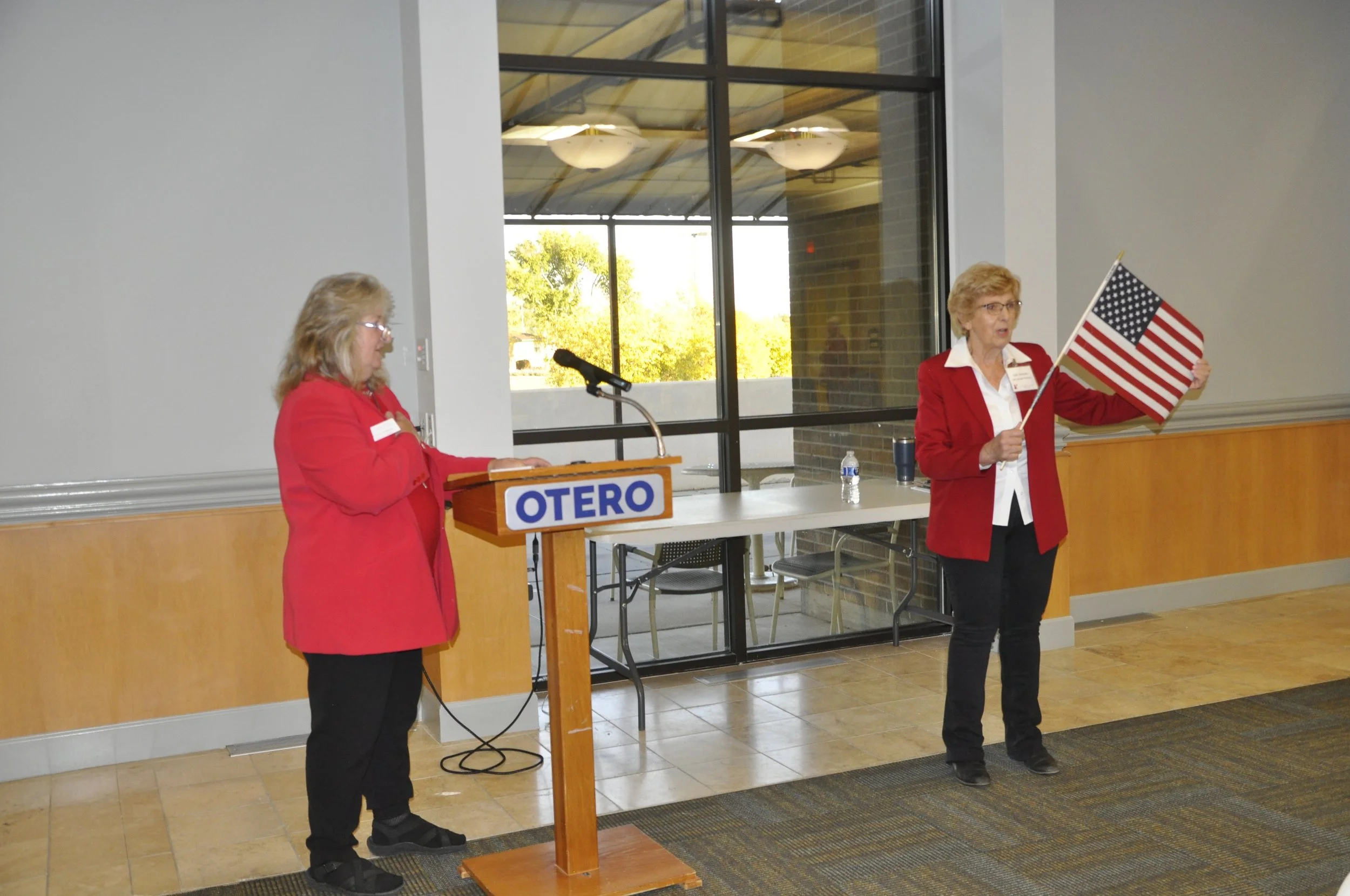 Two women in red blazers participating in an event with a table and a podium labeled 'OTERO'. One woman stands behind a podium, while the other holds a small American flag and gestures with her hand.